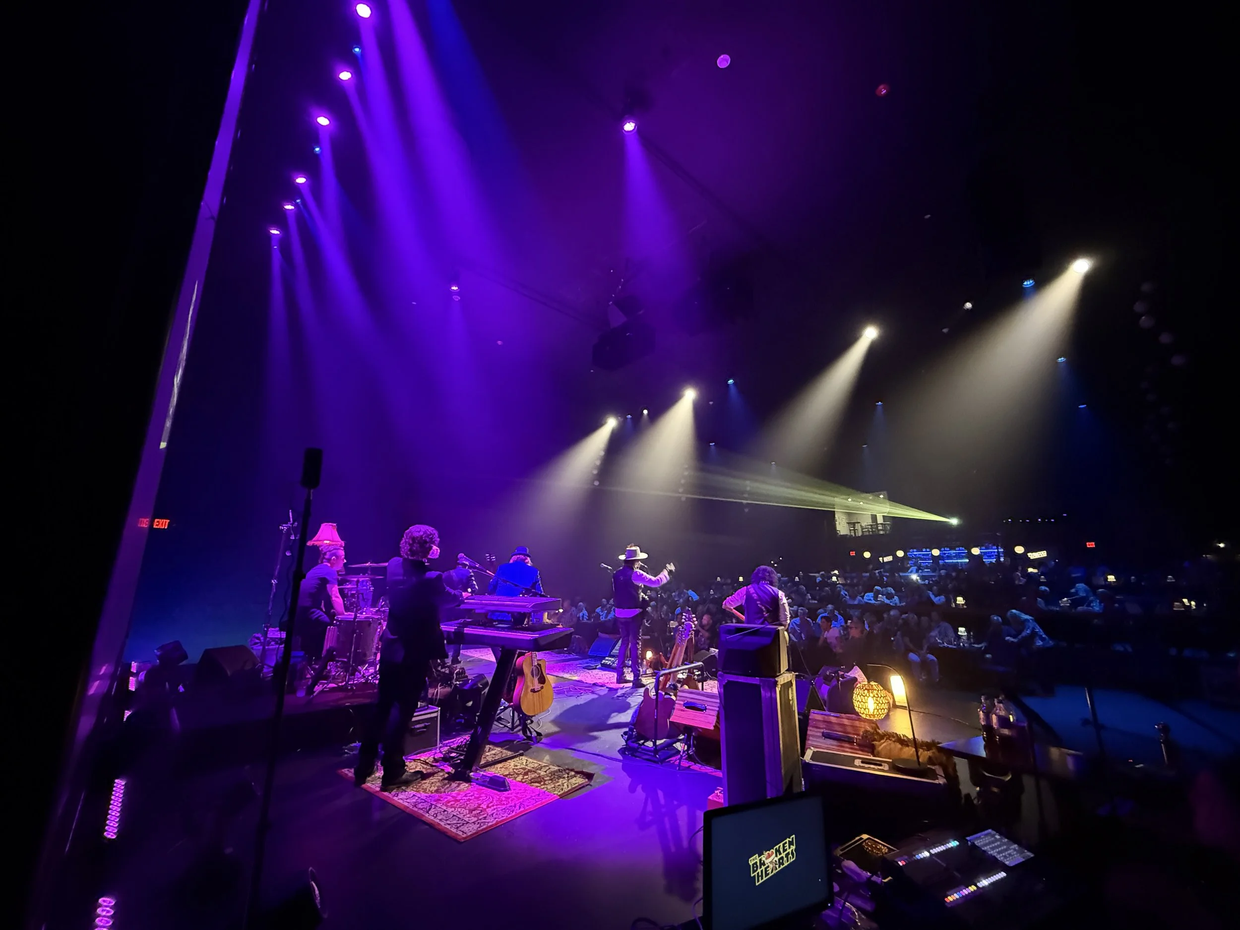 Stage scene with musicians performing under purple and yellow spotlights, audience in the background, with musical instruments and equipment visible. The Lyric Theatre, Blue Island,IL. Chrome Horse: The Bob Dylan Tribute