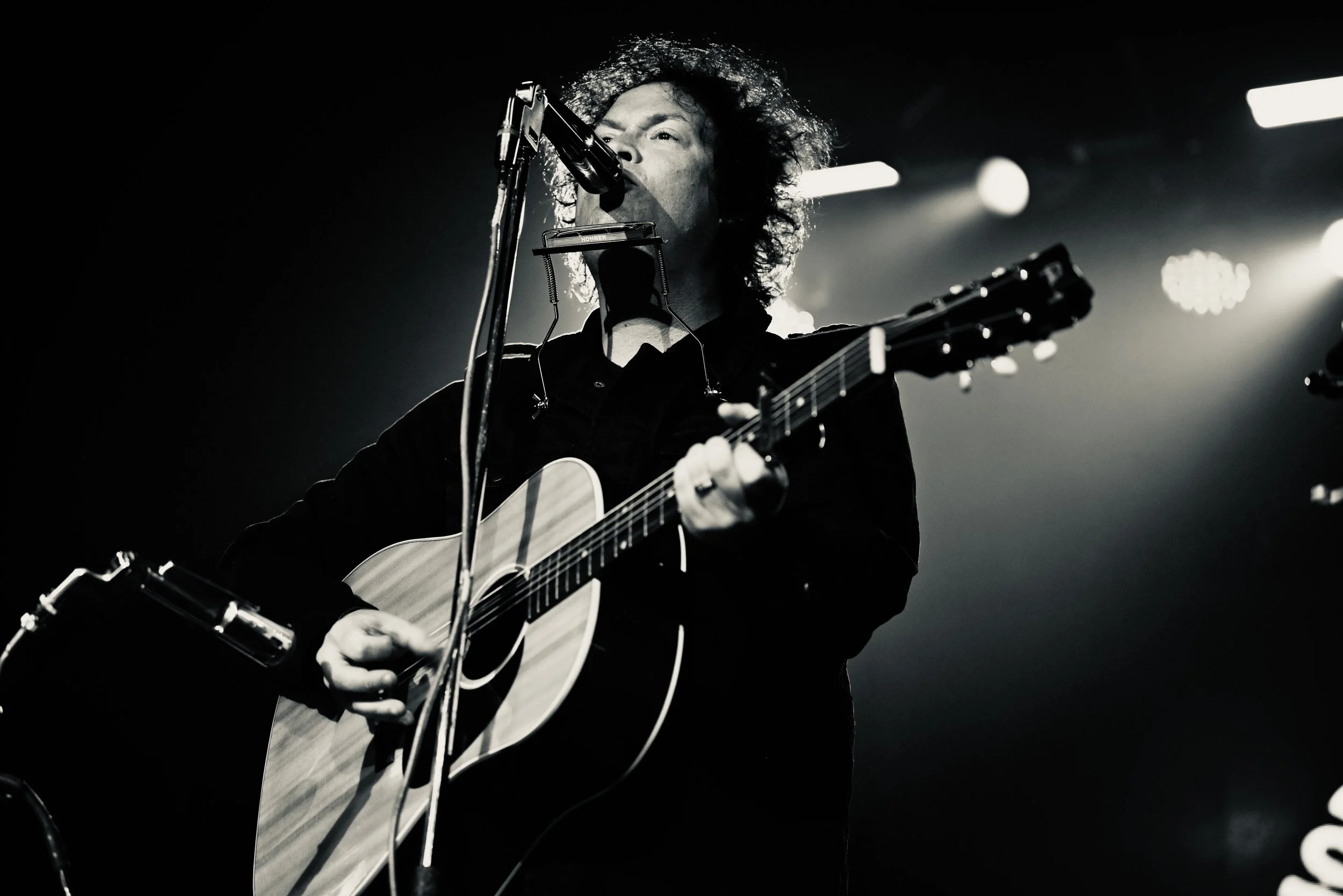 Black and white photo of a male musician with curly hair playing guitar and singing into a microphone on stage with bright spotlights.