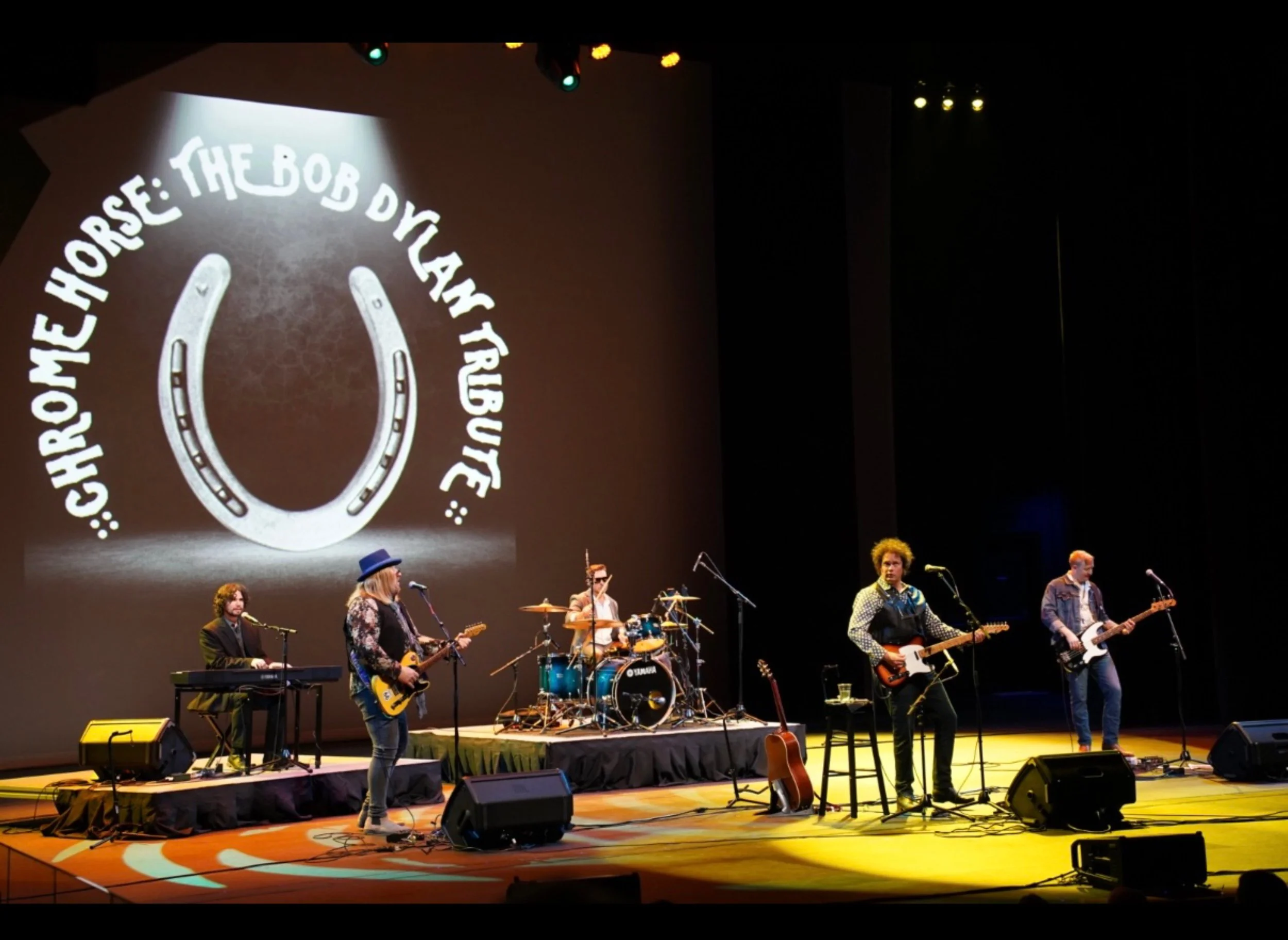 A musical band performing on stage with a large screen behind them displaying the logo of 'Chrome Horse: The Bob Dylan Tribute'. The band has five members, including a keyboardist, a guitarist with long hair and a hat, a drummer, and two other guitar