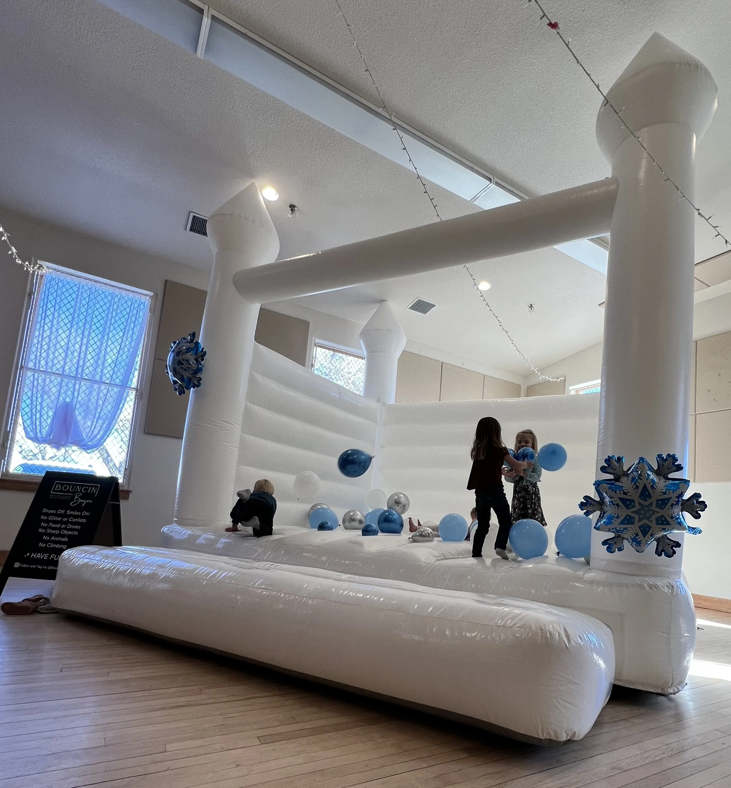 Children playing on an inflatable bounce house decorated with blue, silver, and white balloons, with snowflake accents in an indoor setting.