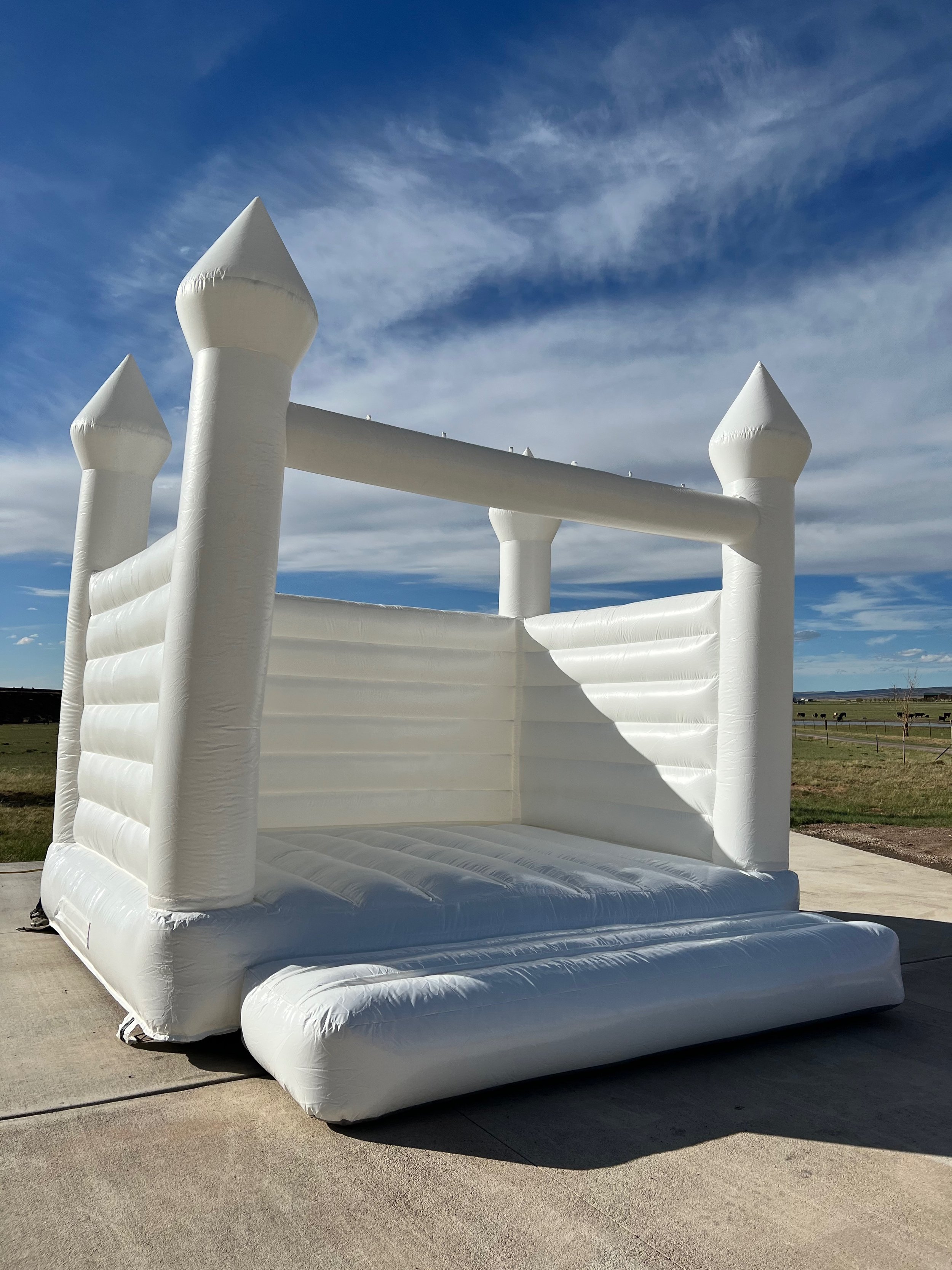 White inflatable bounce house designed to look like a castle with towers at each corner, set outdoors on a concrete surface with a grassy field and blue sky with clouds in the background.