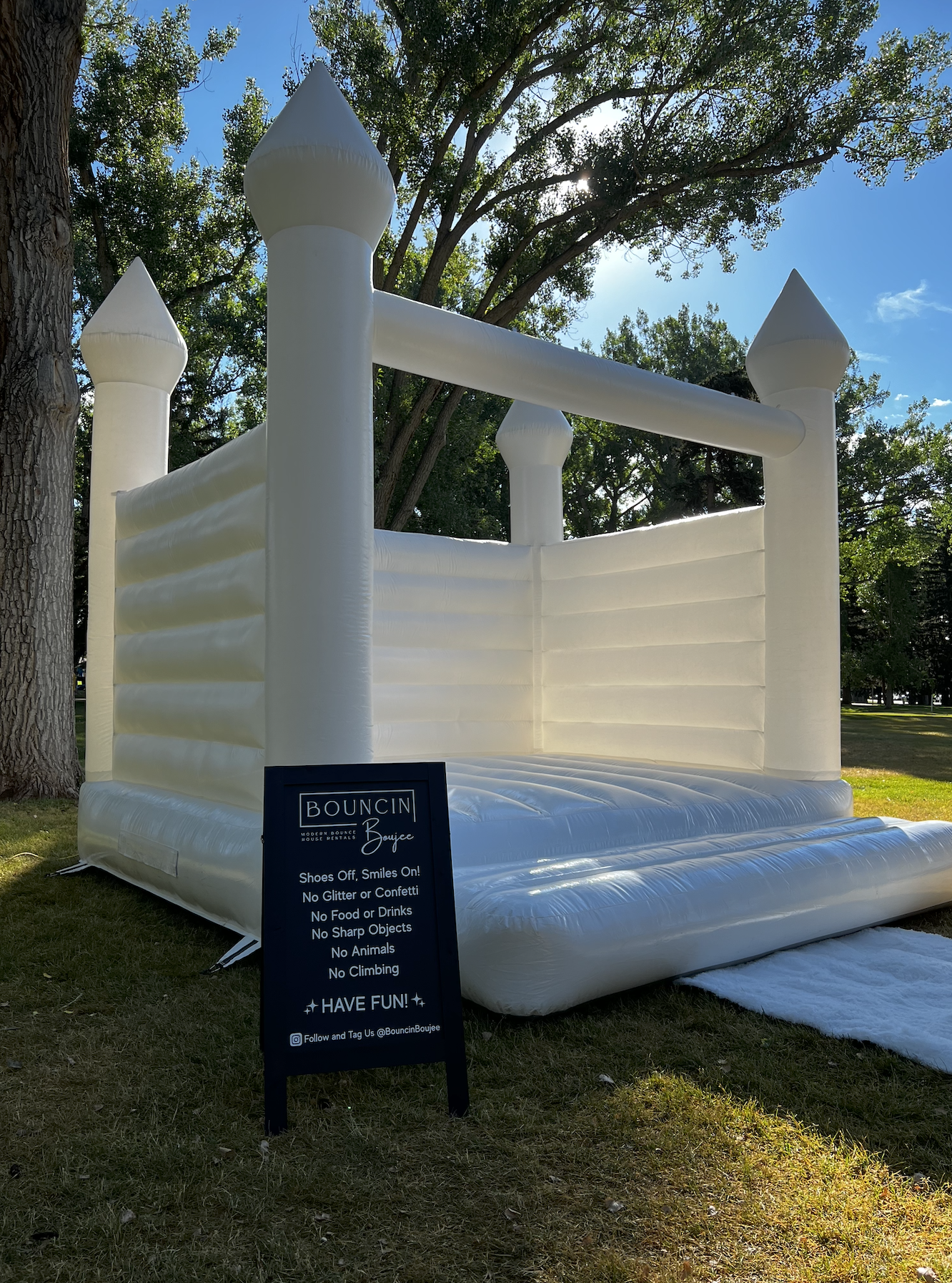 Inflatable white bounce house shaped like a castle with four turrets, set outdoors on grass with trees in the background, and a black sign with rules for use.