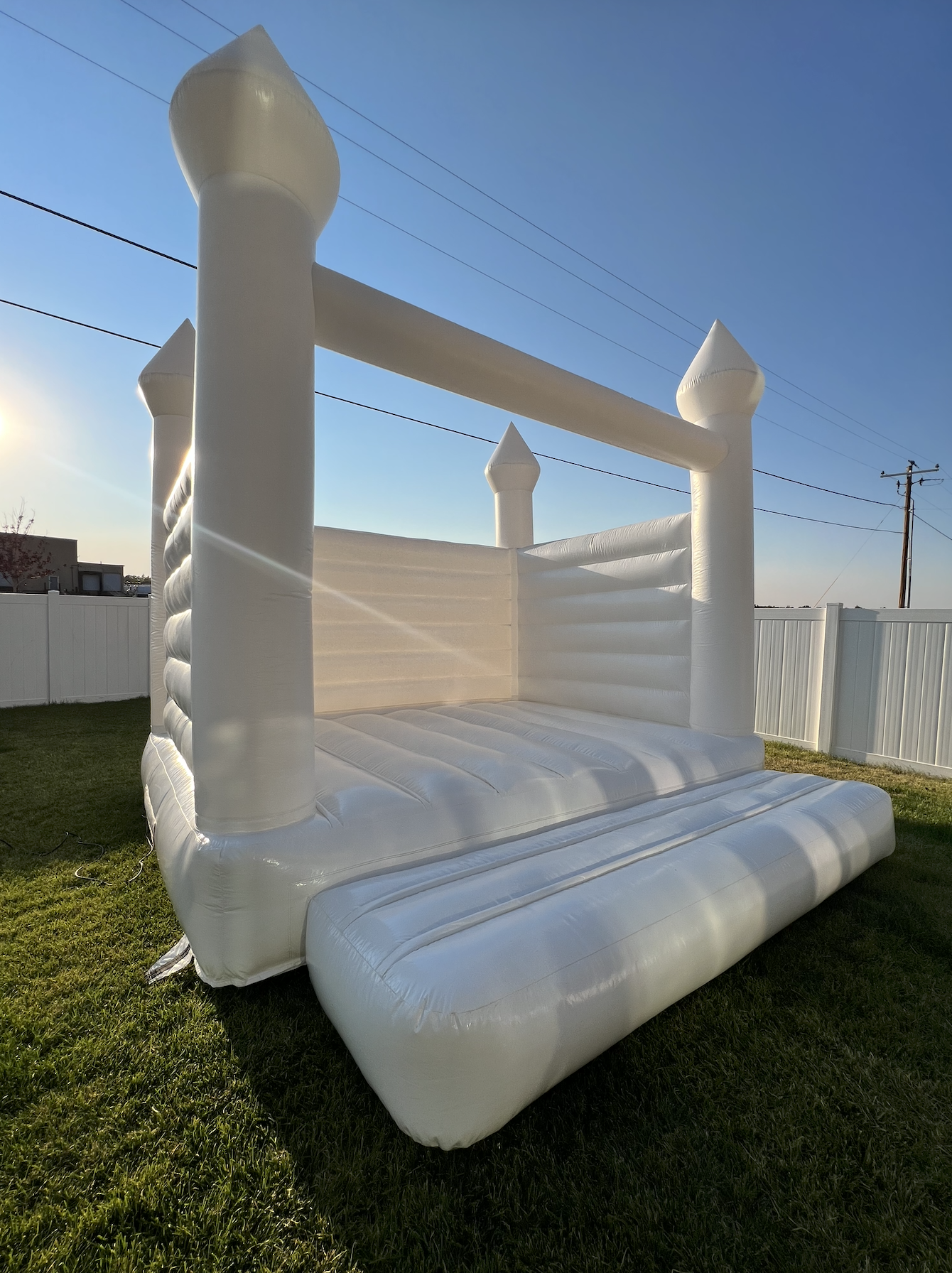 White inflatable bounce house styled like a castle with four towers, set up on a grassy yard with a white fence, under a clear blue sky with sunlight.