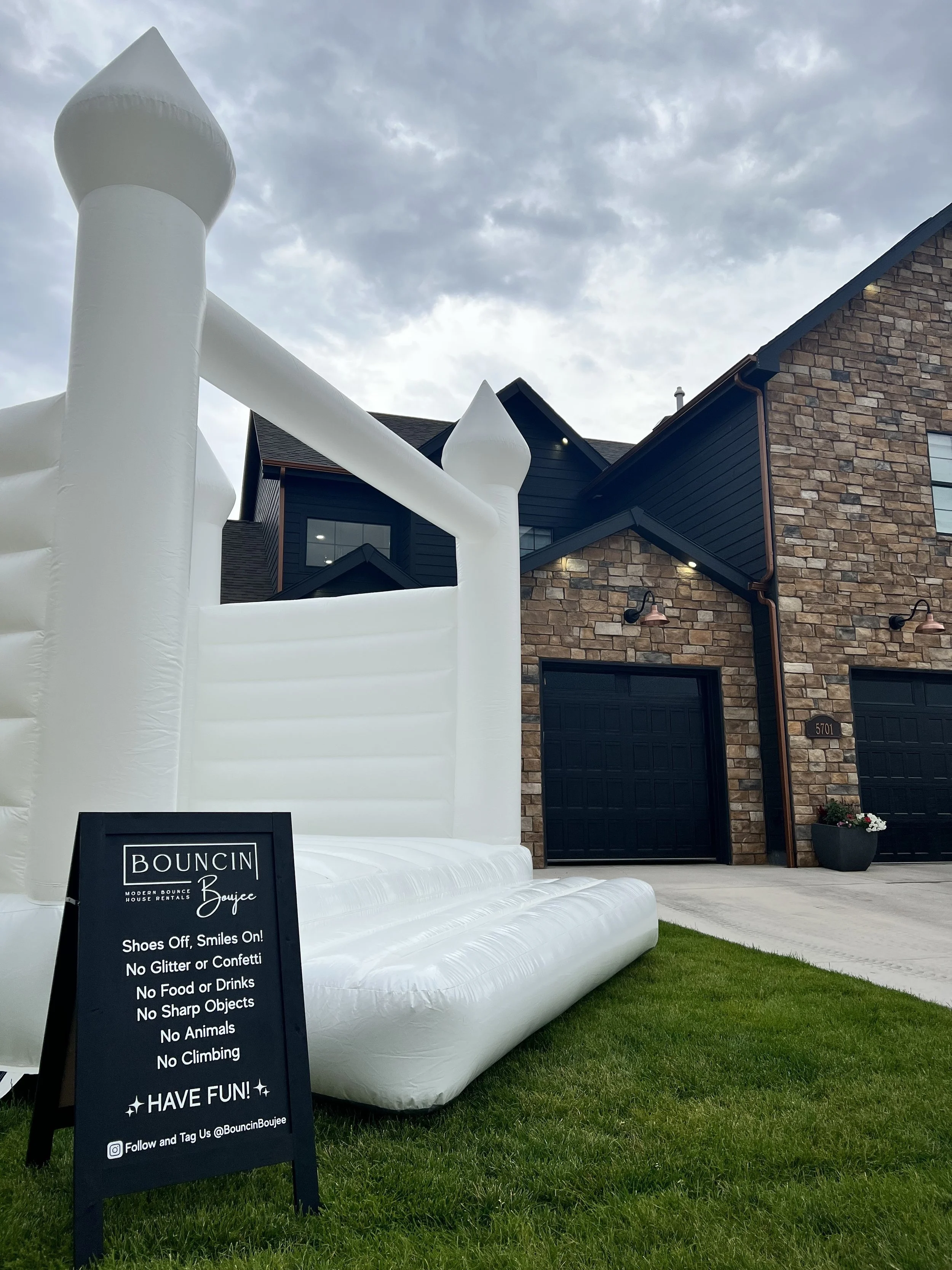 Inflatable bounce house slide in front of a brick house with black garage doors, a flower pot, and a sign with rules for the bounce house, including no shoes, food, drinks, glitter, confetti, sharp objects, animals, or climbing.