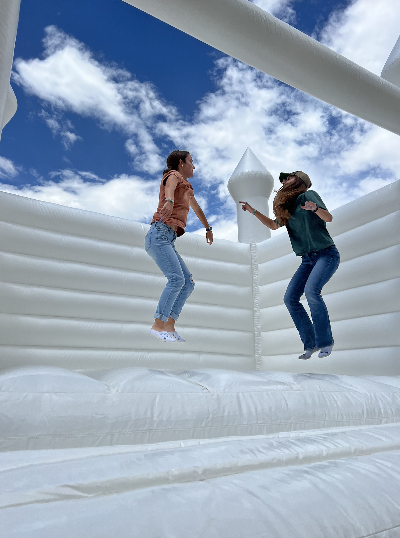 Two women are jumping inside a white inflatable structure with a blue sky and clouds in the background.