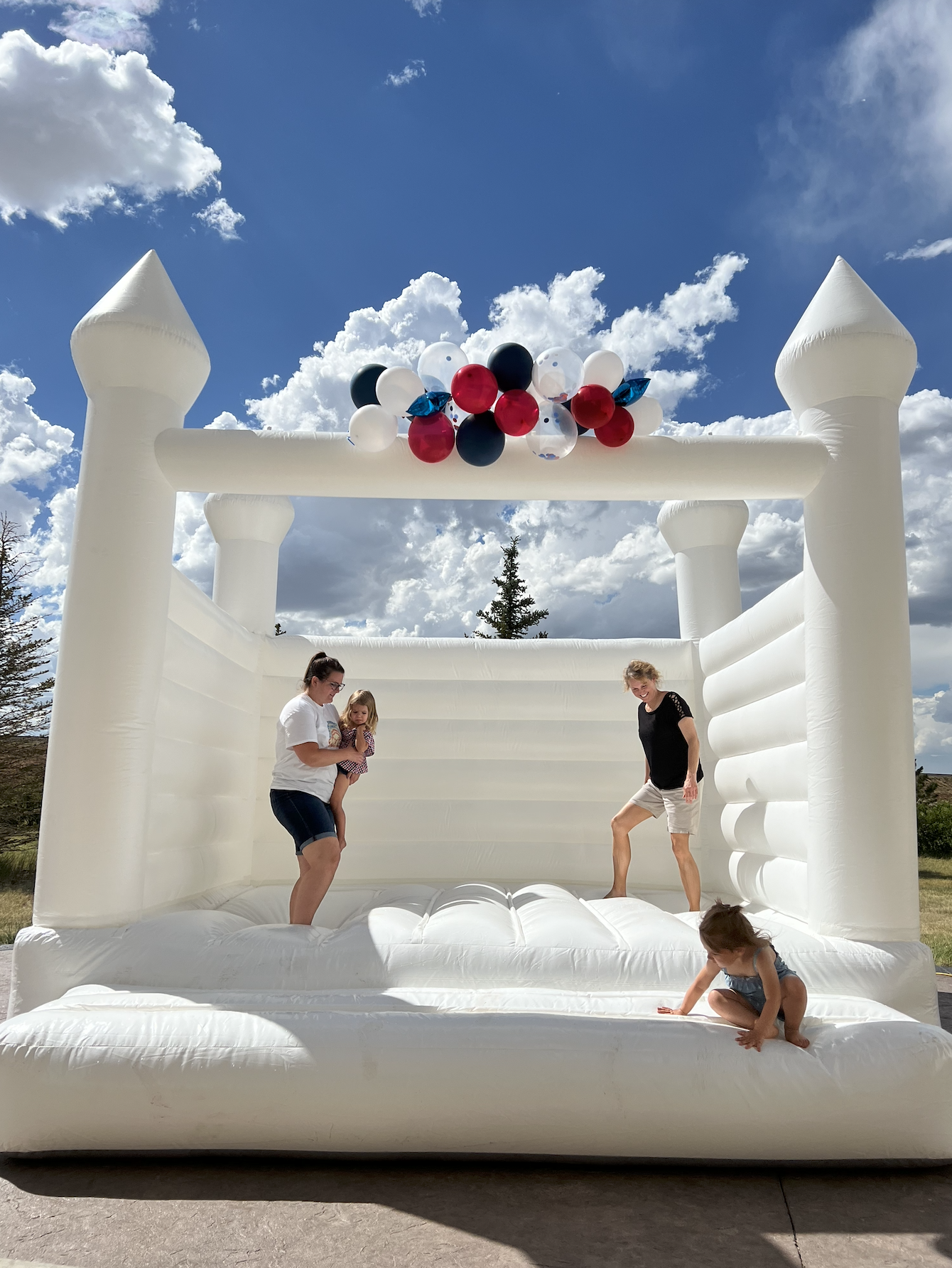 People playing inside a white inflatable castle decorated with balloons, outdoors on a sunny day with a partly cloudy sky.