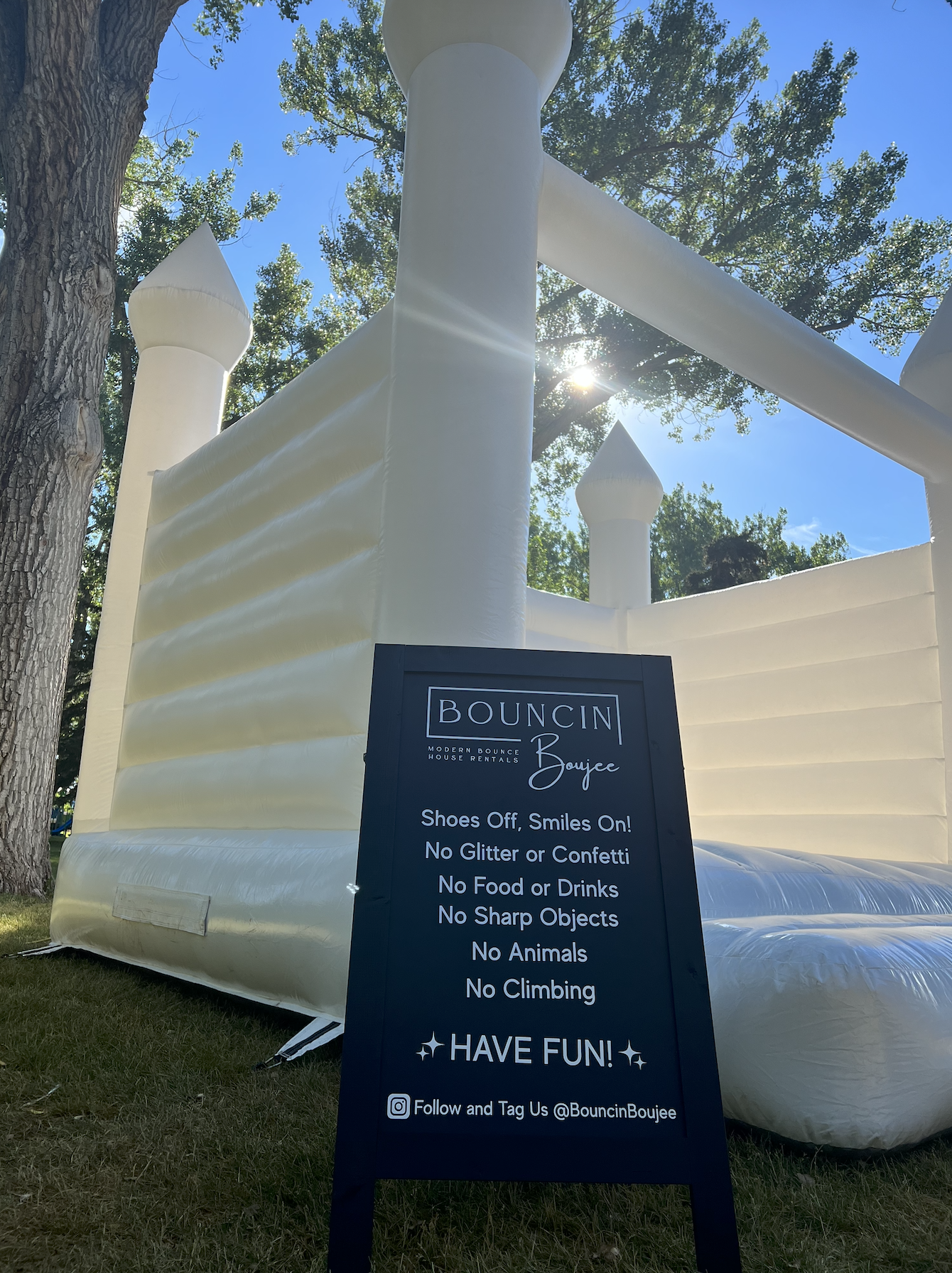 A white inflatable bounce house with turret-like structures, set outdoors on grass, with a sign in front listing rules for jumping, including no shoes, glitter, alcohol, sharp objects, animals, or climbing, and encouraging fun, during daytime with bl