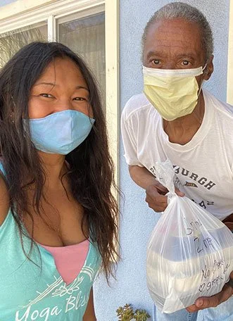 A young woman and an older man standing outside, both wearing face masks. The woman is smiling, and the man is holding a plastic bag.