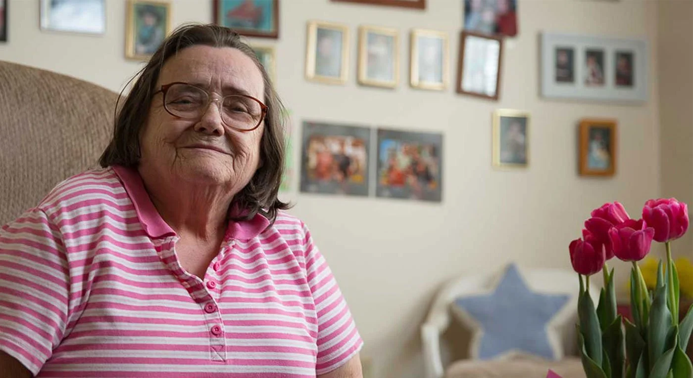 An elderly woman with glasses wearing a pink and white striped shirt, smiling softly, sitting in a room with framed photographs on the wall and a vase of pink tulips in the foreground.