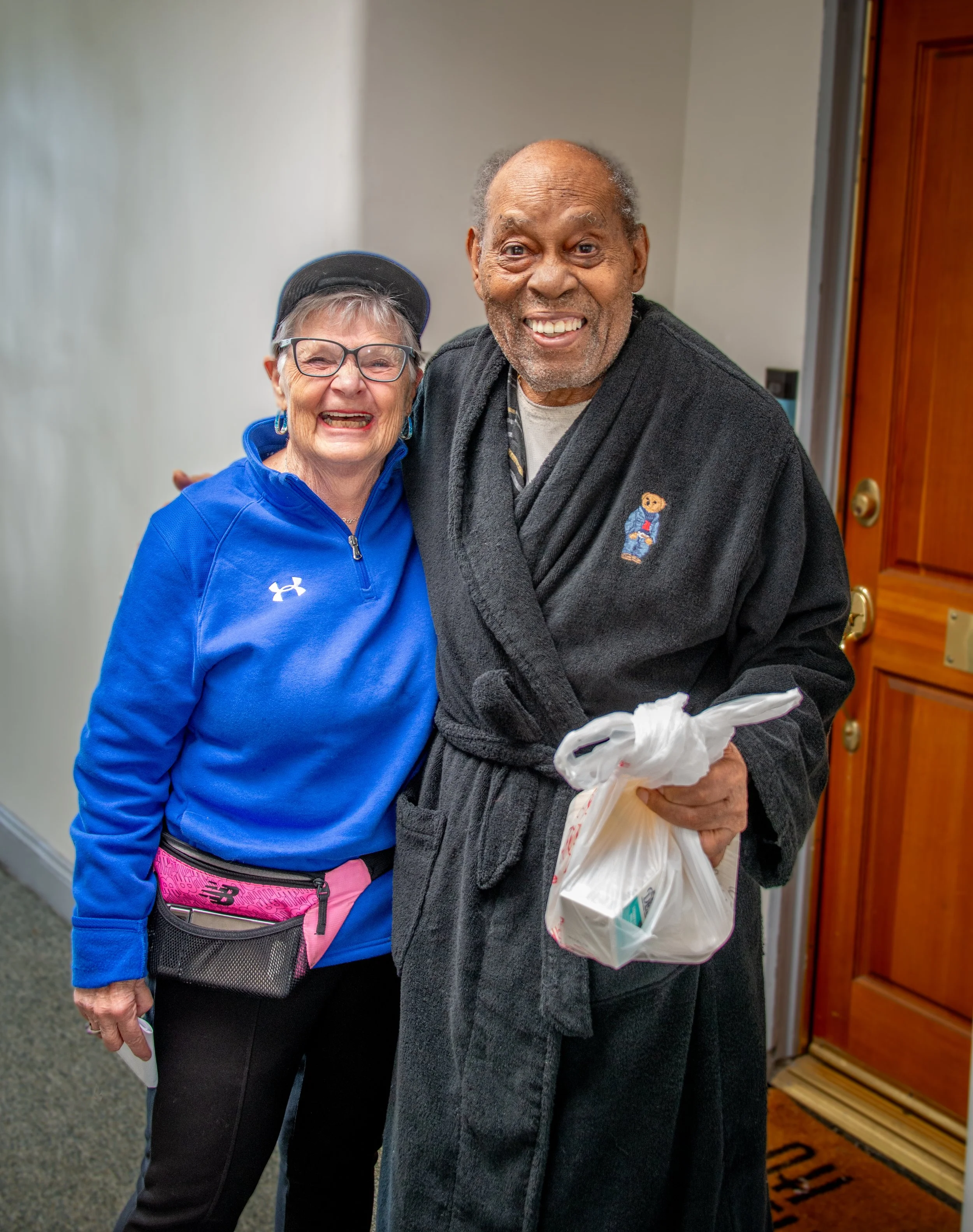 An elderly woman in glasses and a blue jacket standing next to an elderly man in a gray robe, holding a white plastic bag, both smiling warmly.