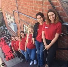 A group of six women standing on a staircase outdoors, all wearing matching red shirts, with a brick wall behind them.