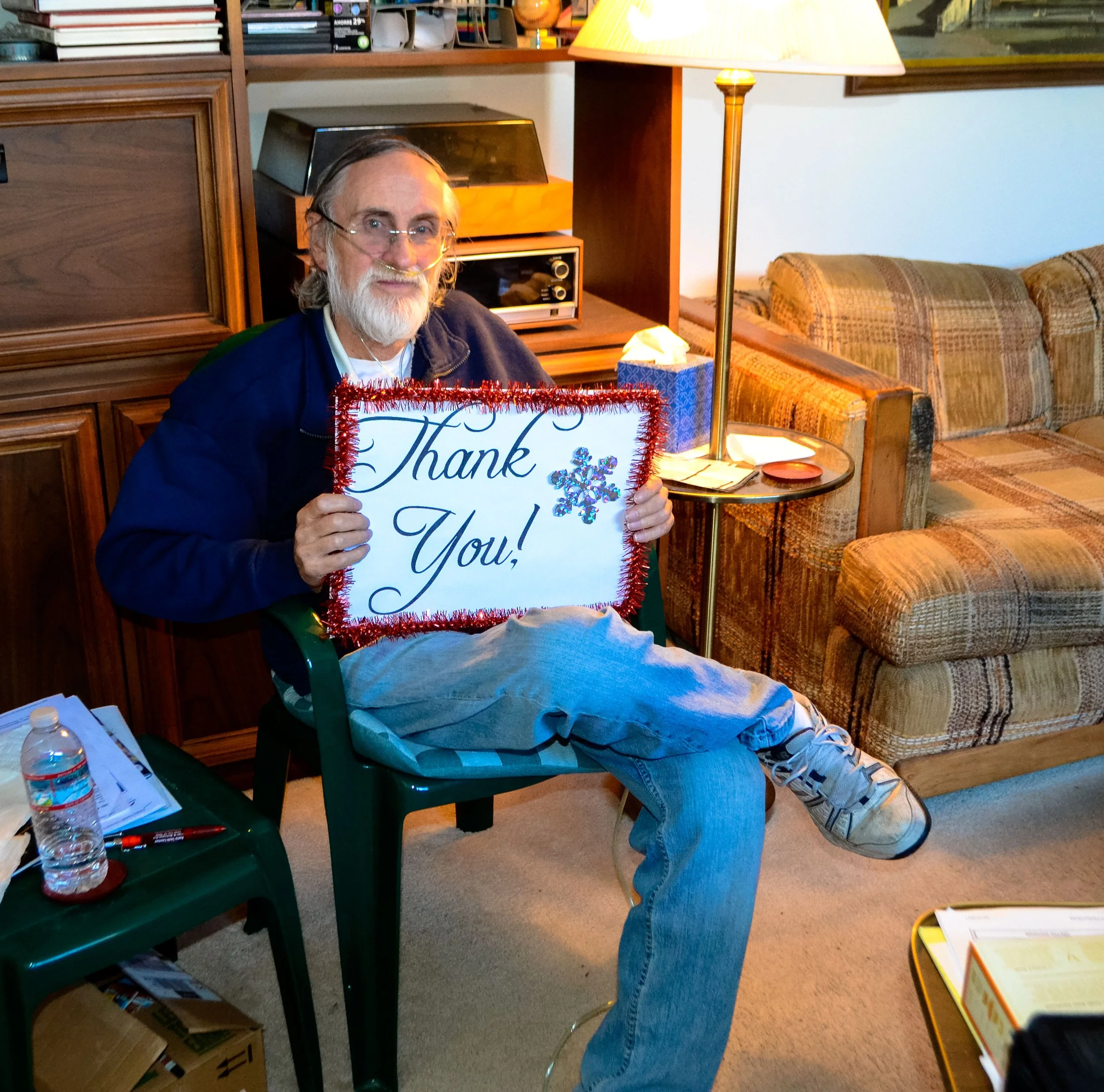 Someone sitting in a living room holding a 'Thank You' sign decorated with tinsel and snowflakes. The person has a beard and glasses, and is wearing a jacket and jeans. The room has a patterned sofa, a table with tissues, and a lamp.