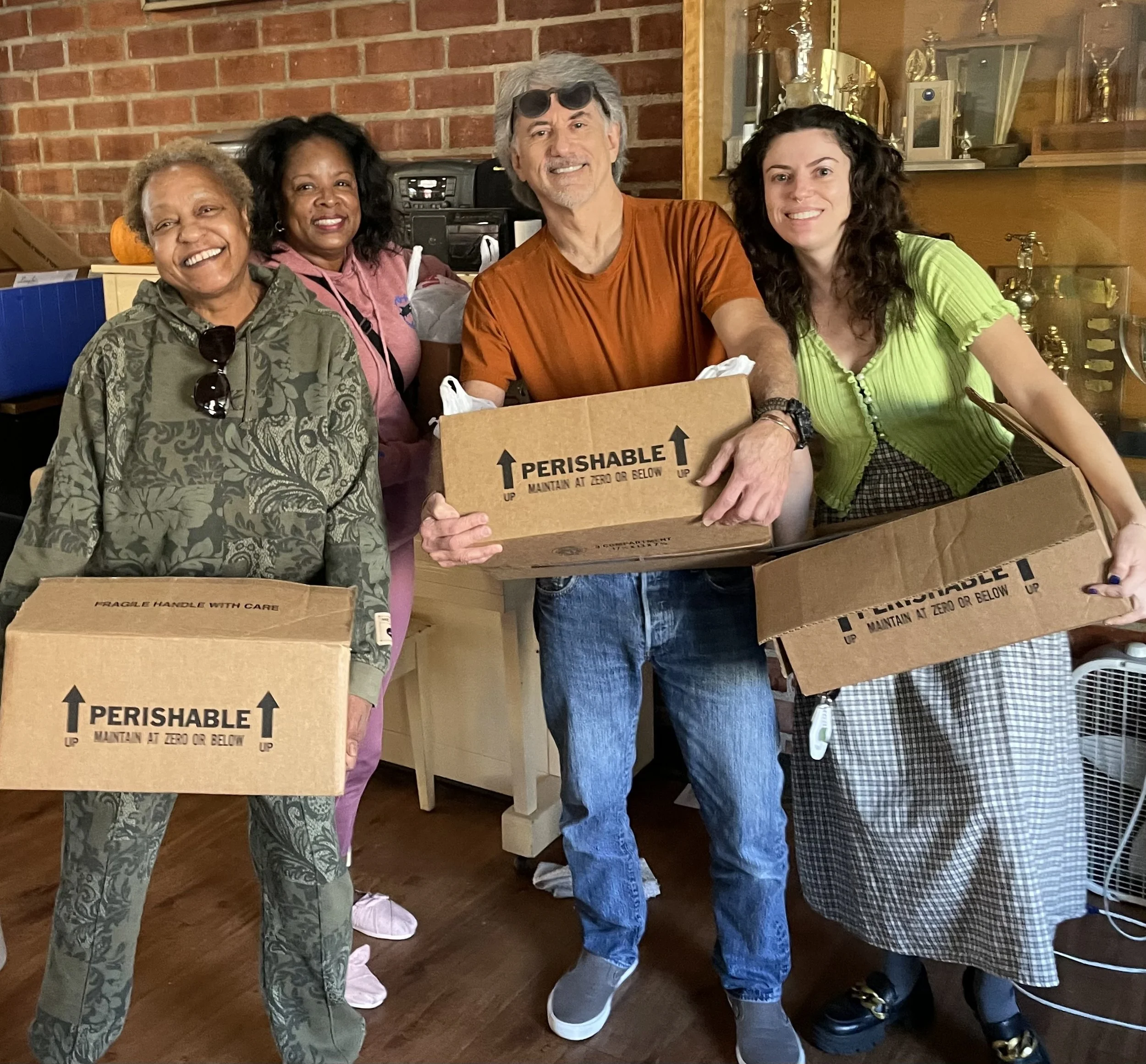 Group of five people smiling and holding cardboard boxes in a room with brick walls and wooden furniture.
