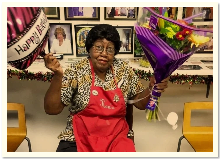 An elderly woman wearing glasses and a leopard print blouse holding a bouquet of flowers and a balloon with a birthday message, sitting in front of a decorated wall.