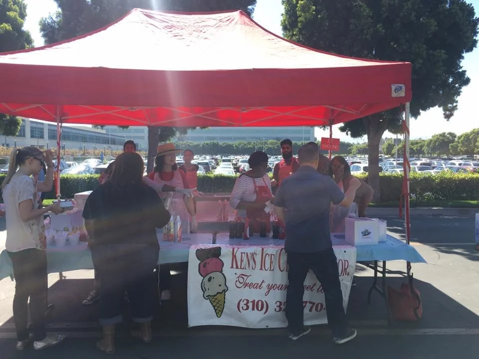 People gathered around a red tent at an outdoor ice cream stand with a banner advertising ice cream flavors, some talking and purchasing ice cream.