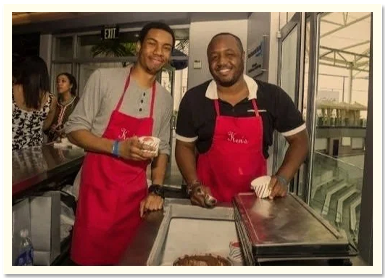 Two men wearing red aprons standing behind a counter at a food stall or cafe, smiling at the camera. One is holding a food item, with a cake in front of them, and there are other people and large windows in the background.