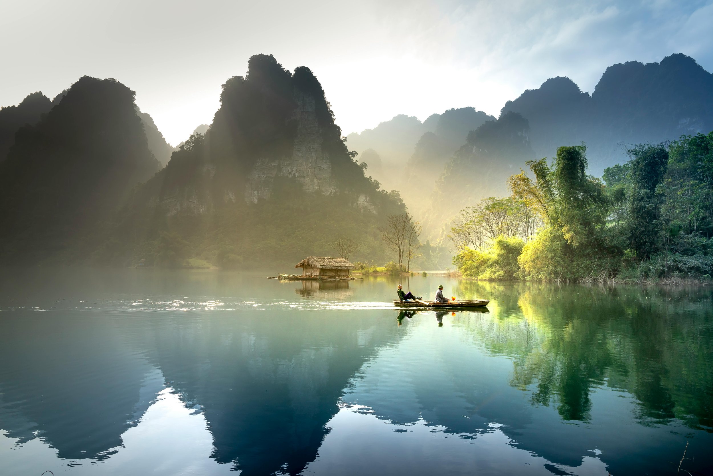Two people in a small boat on a calm river with lush green trees and towering mountains in the background, during sunrise or sunset.