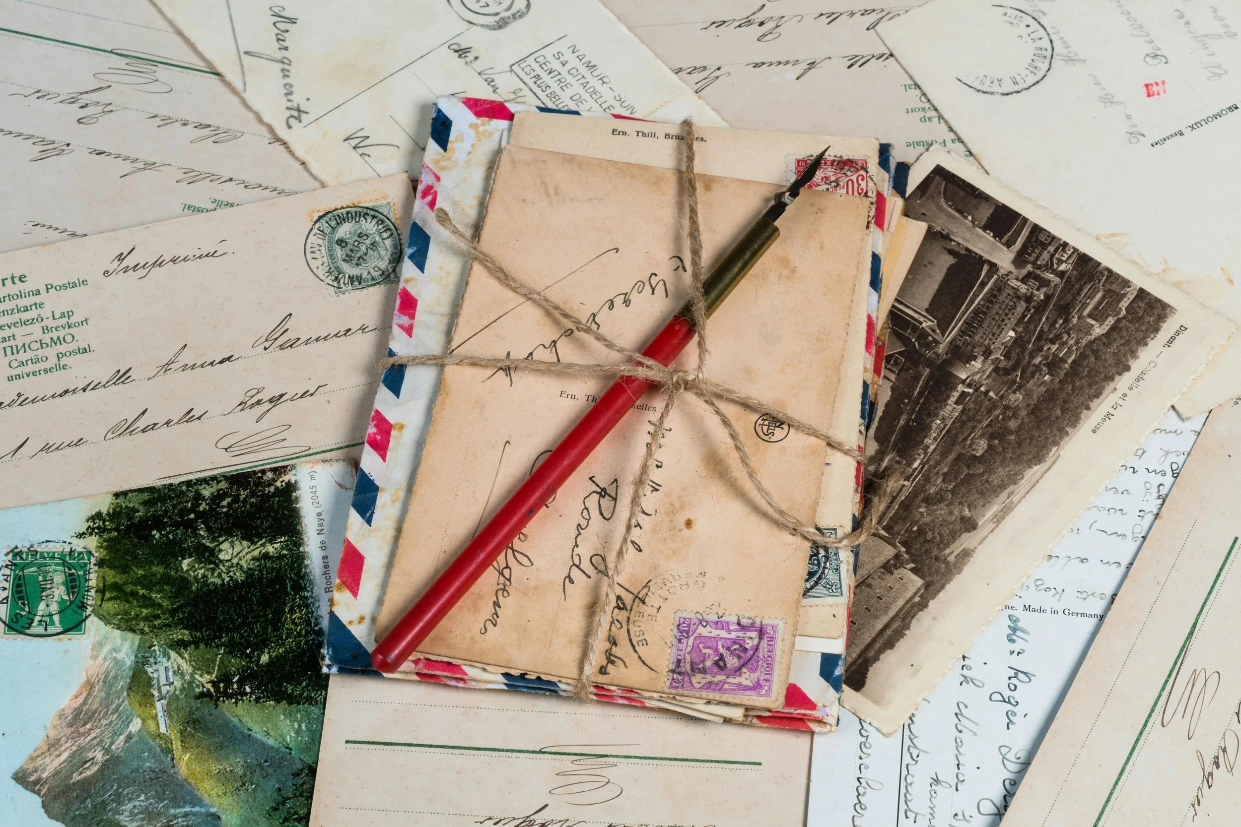 A collection of vintage postcards and letters, with one tied with string, a fountain pen resting on top, and some seen in black and white and colored.