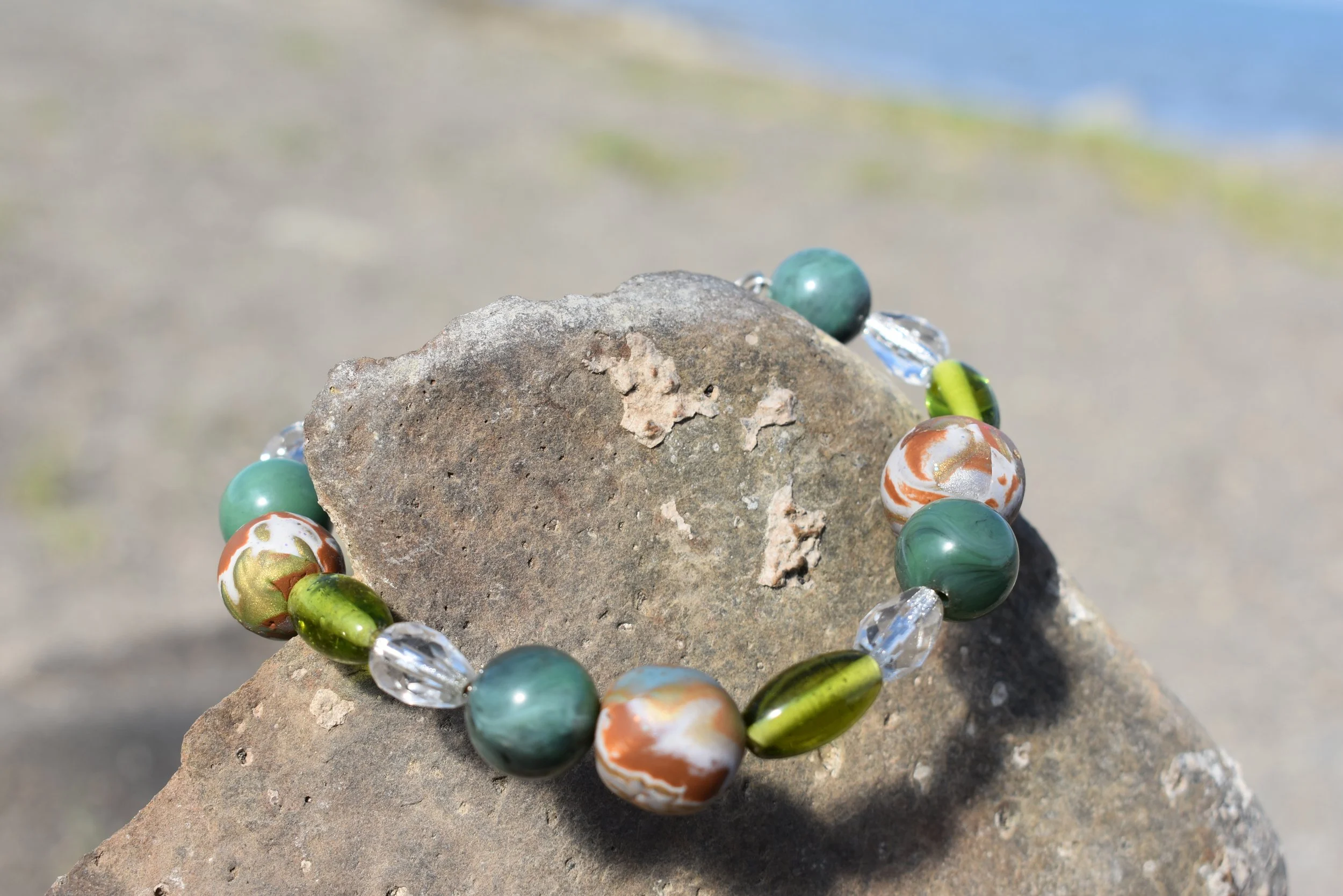 A colorful bead bracelet resting on a large rock at the beach with sand and water in the background.
