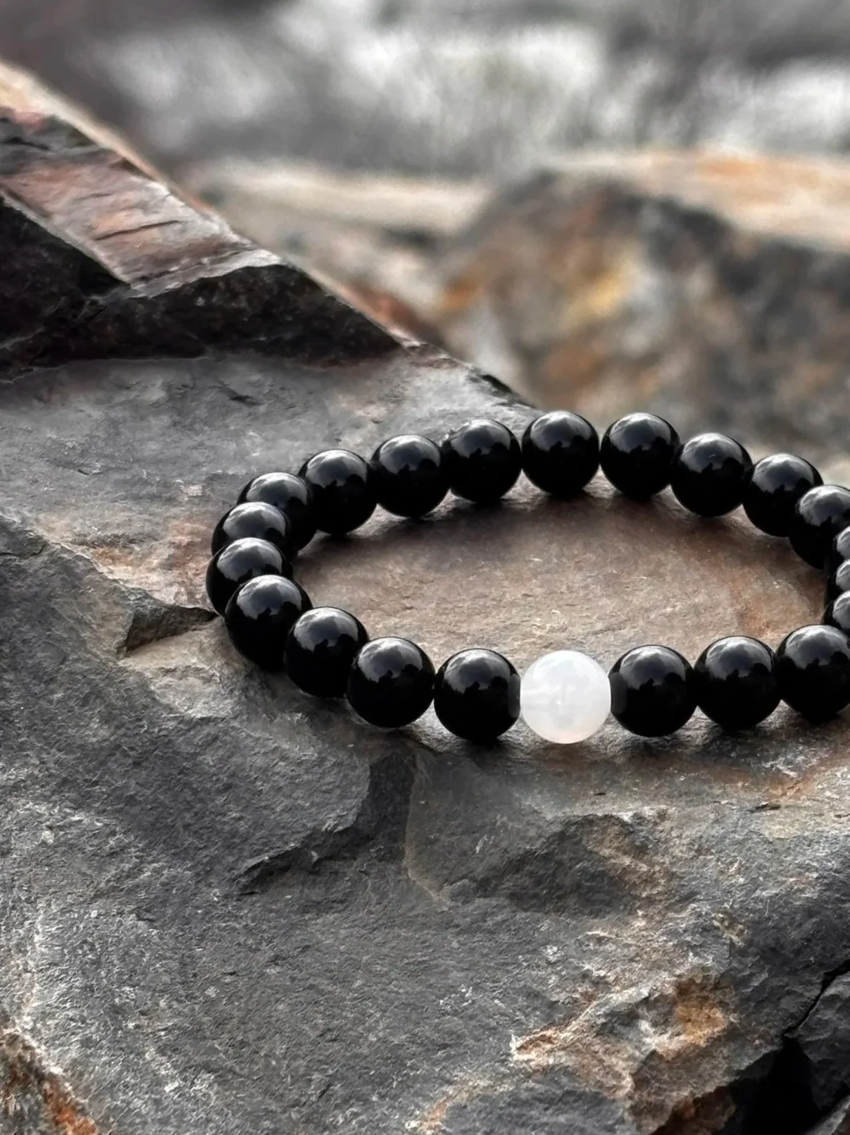 A beaded bracelet featuring black beads with a single white bead, resting on a rough stone surface.