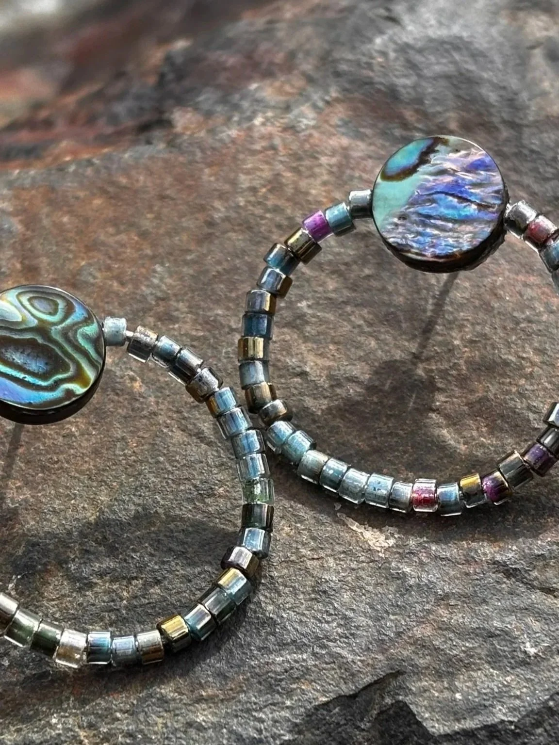 Close-up of beaded hoop earrings with iridescent circular accents resting on a textured stone surface.