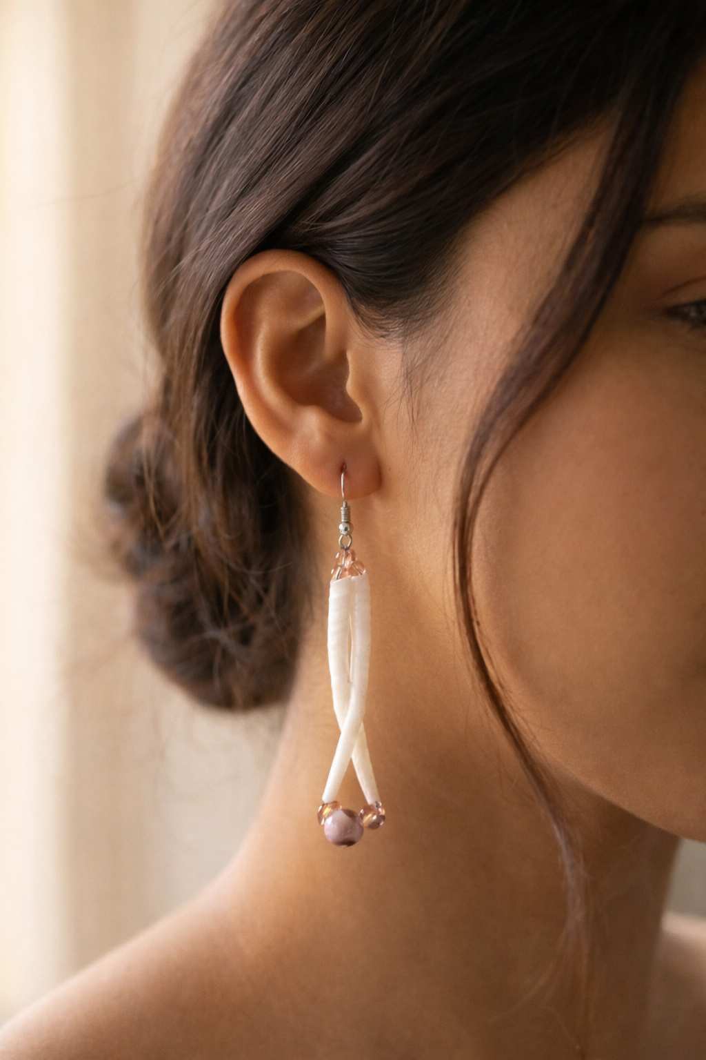 Close-up of a woman's ear wearing a dangling earring with a white, curved, textured piece and small beads at the end.