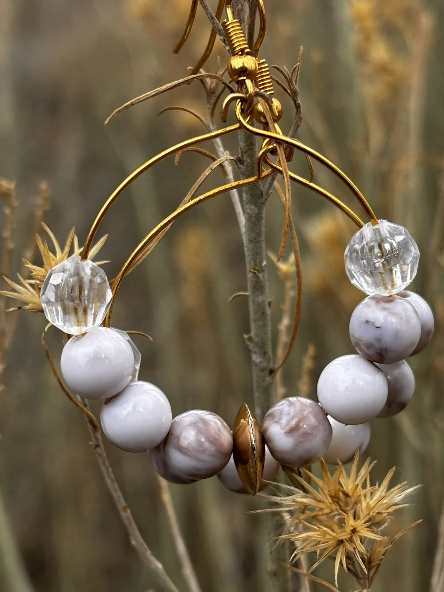 A pair of beaded earrings with gold hooks hanging from a tree branch with dried grass and twigs in the background.