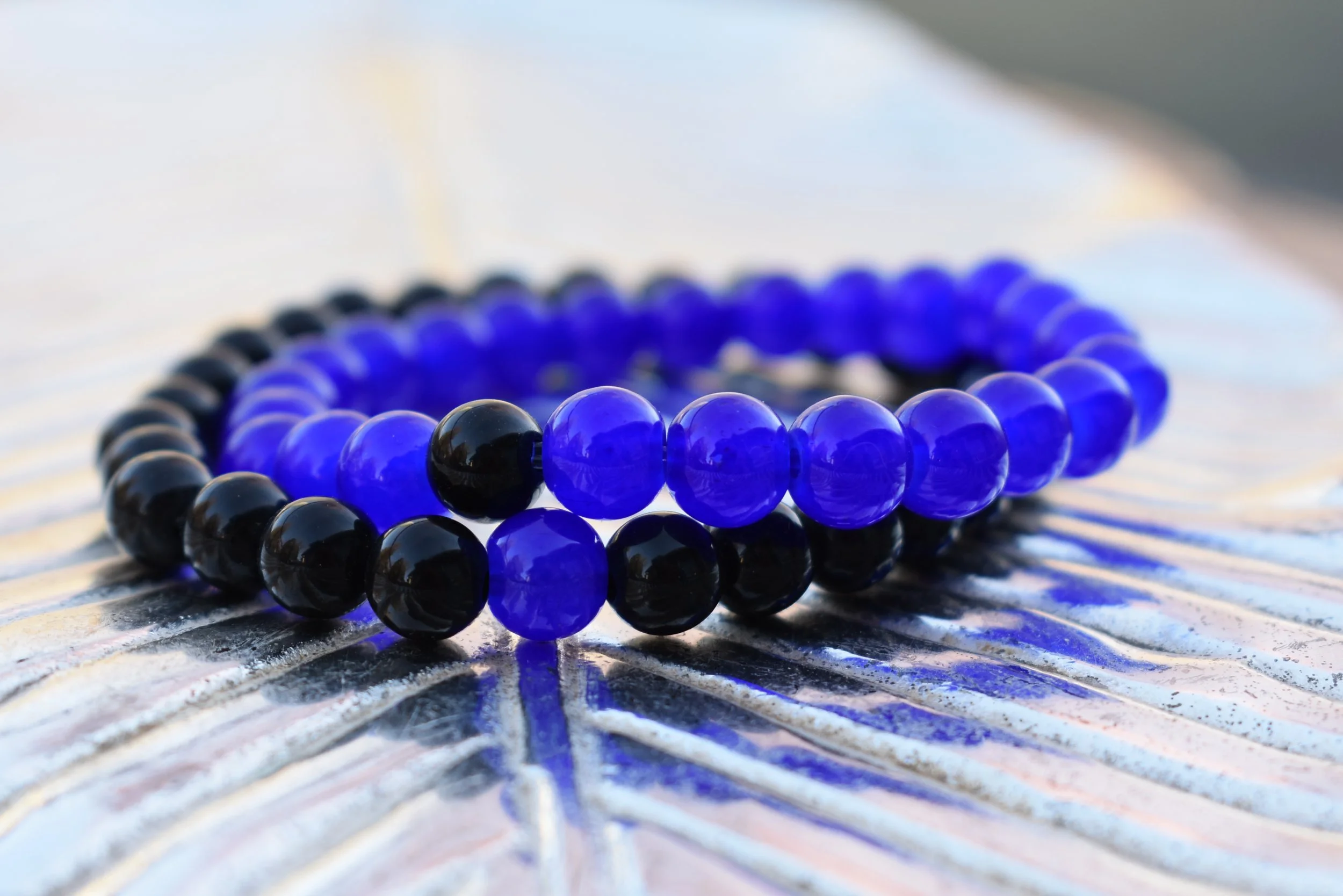 Close-up of two beaded bracelets, one with black beads and the other with blue beads, placed on a textured, iridescent surface.