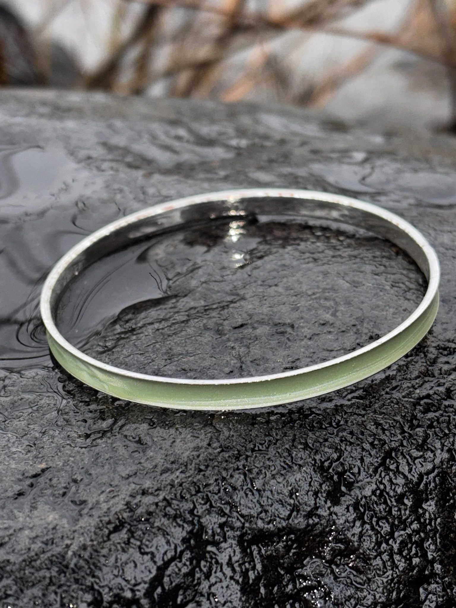 A green and silver metal bracelet resting on wet, black, rough textured surface, with blurred natural background.