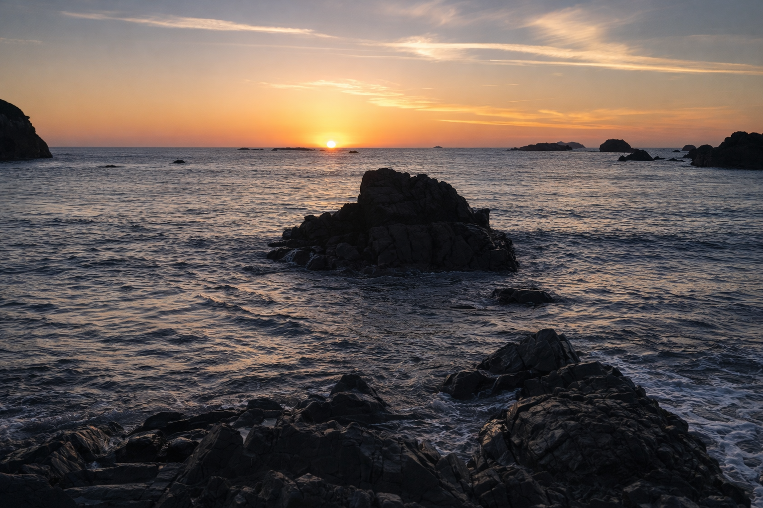 A sunset over the ocean with rocks in the foreground and distant islands on the horizon.