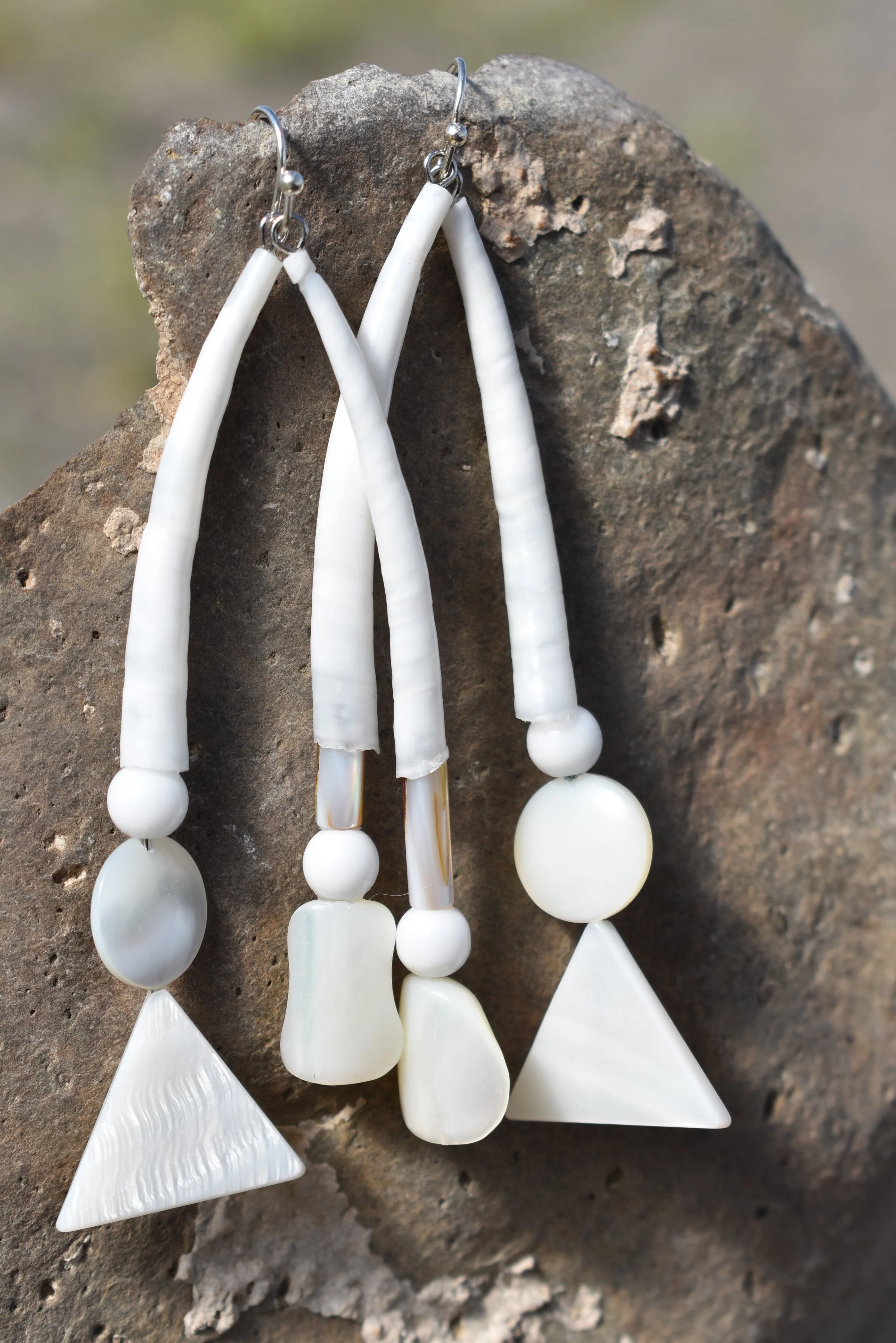 Pair of white, shell-made earrings with various textured beads, hanging on a rough rock surface.