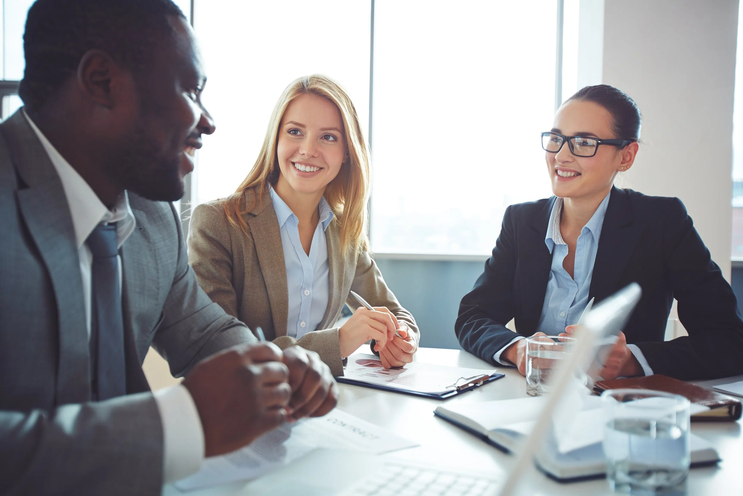 Three business professionals in a meeting room, two women and one man, engaged in conversation with documents and laptops on the table.