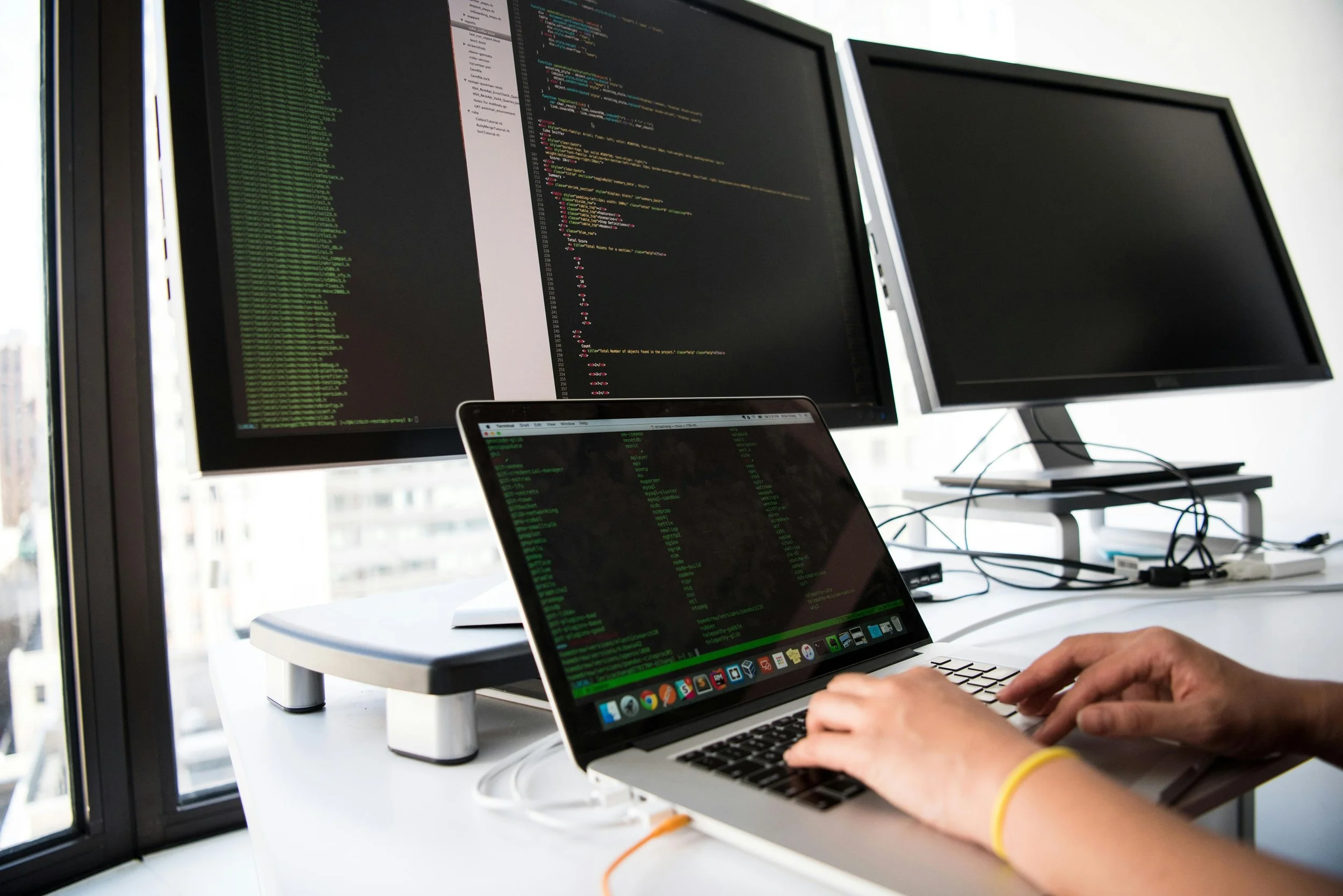 Person working on a laptop with multiple monitors displaying programming code inside an office near large windows.