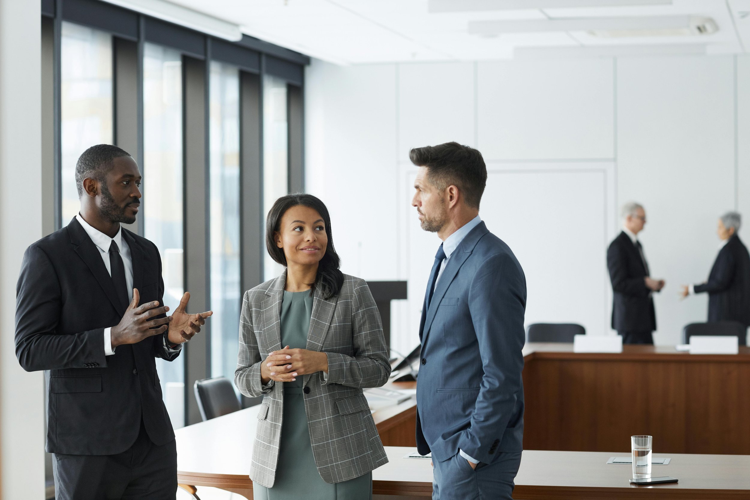 Three business professionals engaging in conversation in a modern office with large windows, in the background two people are having a discussion near a table.