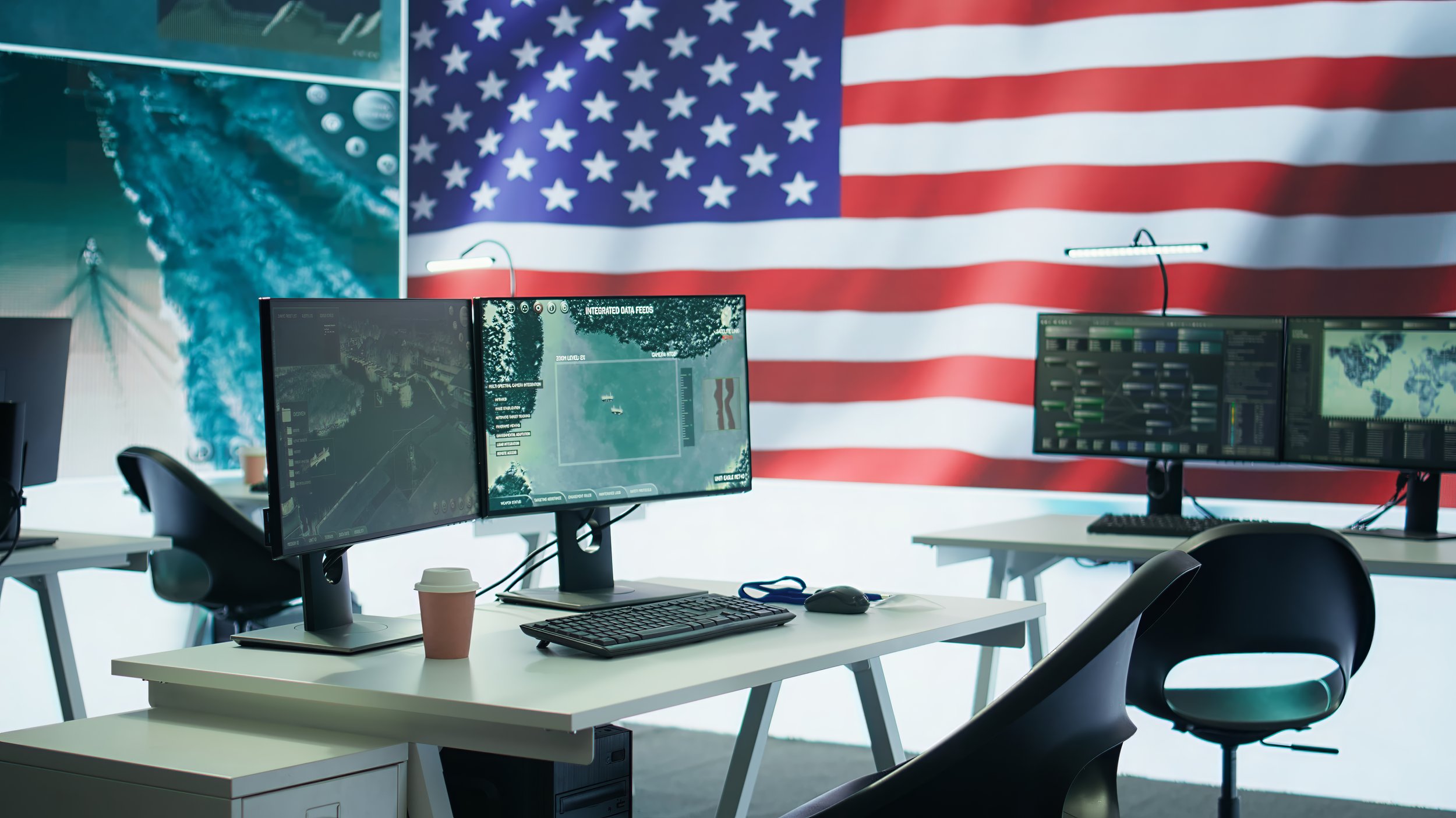Office workspace with dual computer monitors displaying data feeds and maps, a keyboard, mouse, disposable coffee cup, and American flag hanging on the wall in the background.