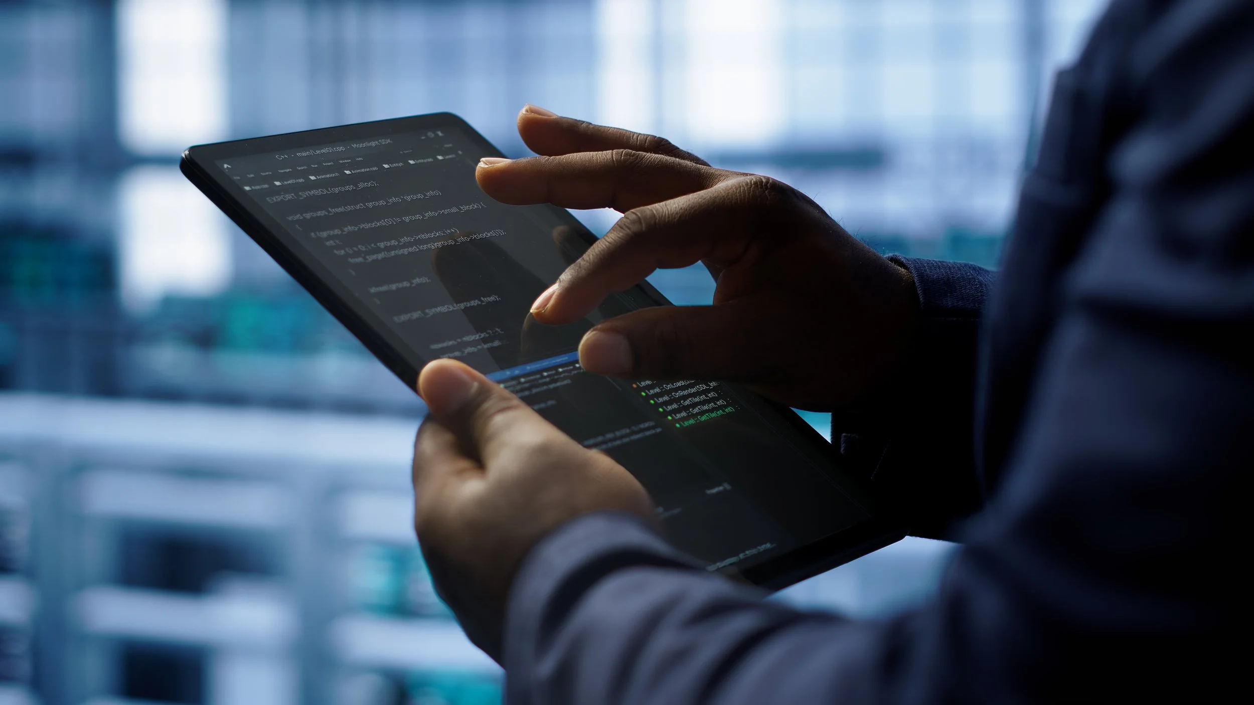 Close-up of a person using a tablet with code on the screen, in a modern office with large windows and a blurred cityscape in the background.