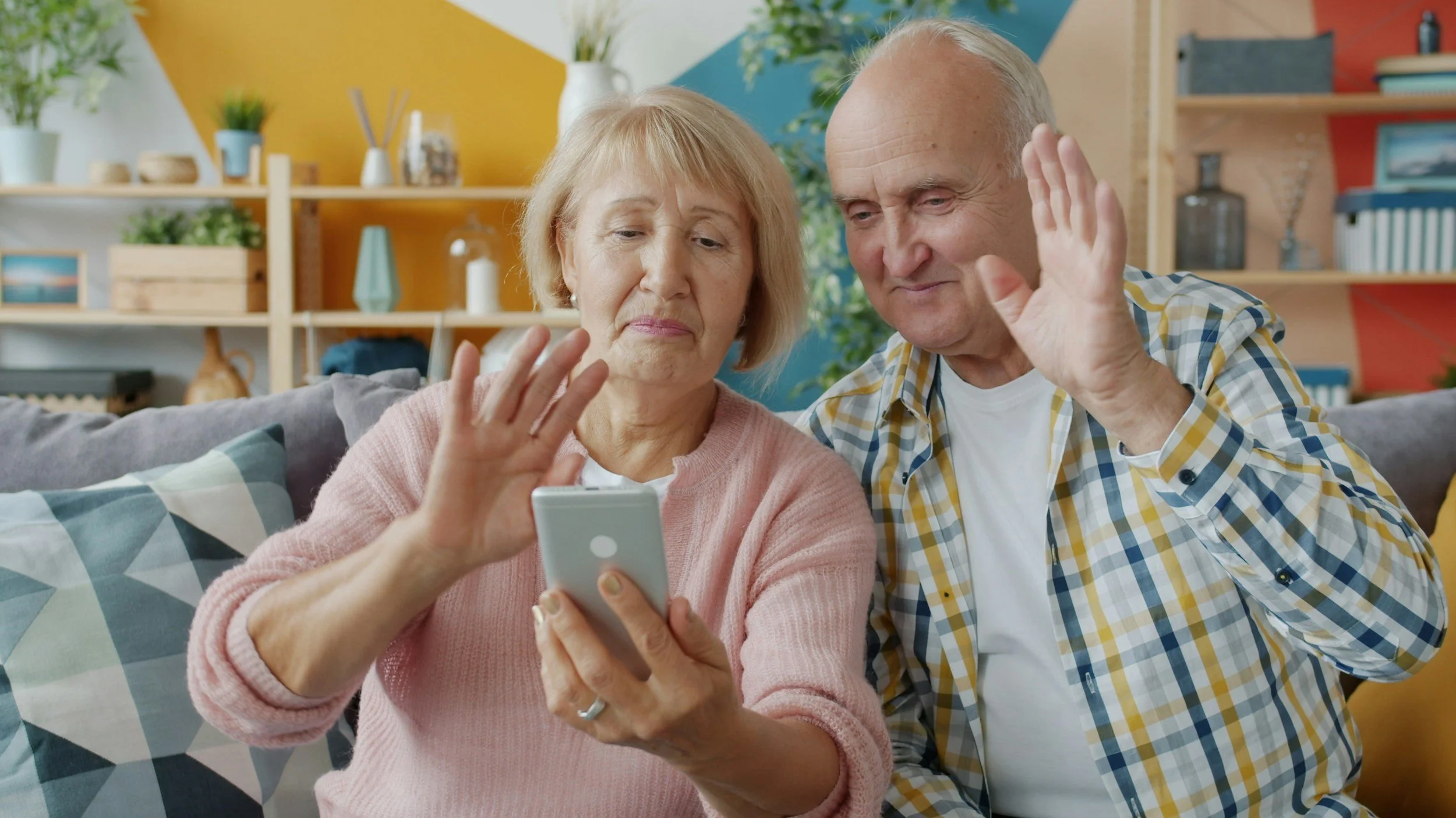 An elderly couple sitting on a couch, looking at a smartphone together, in a colorful and well-decorated living room.