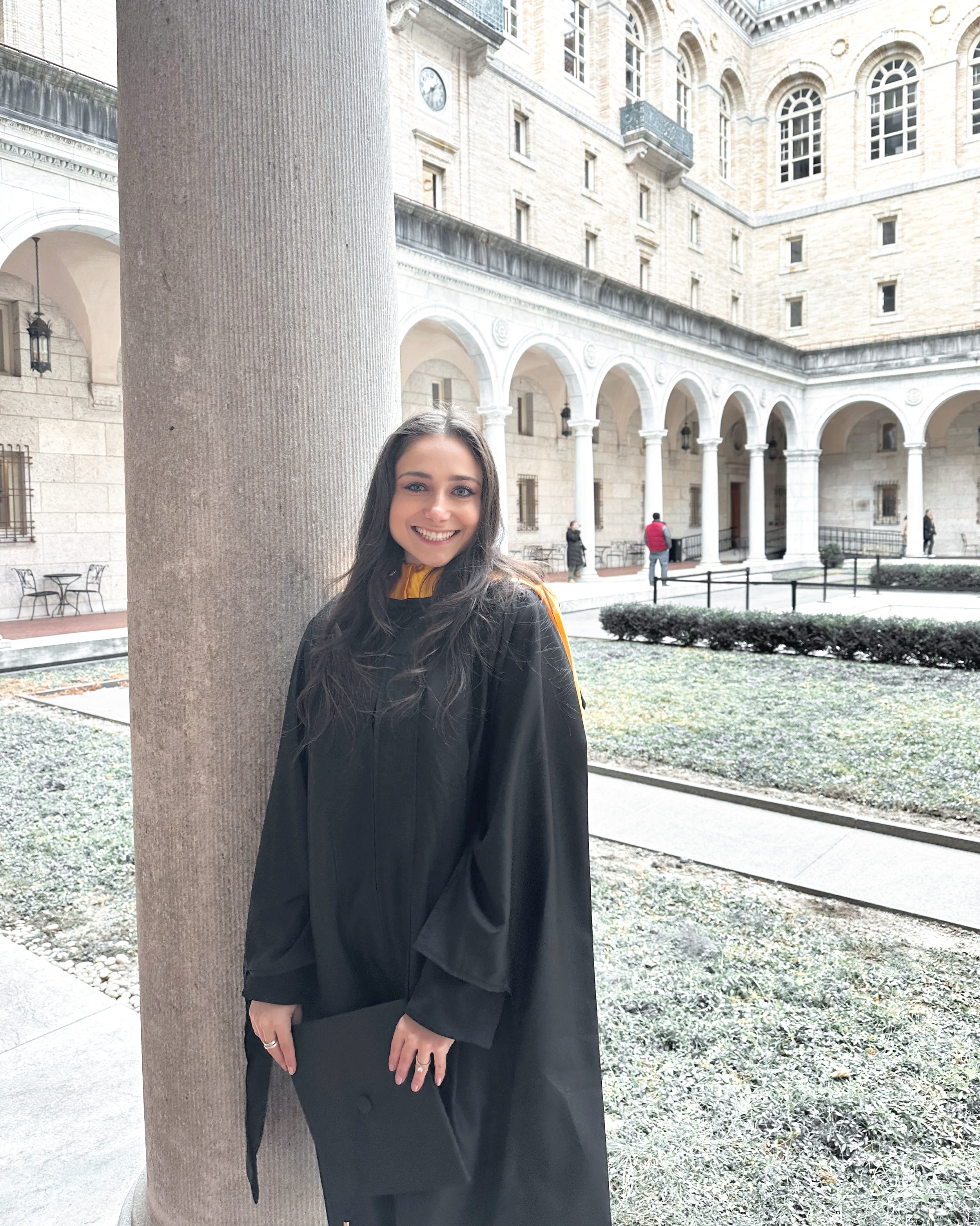 A young woman in a graduation gown holding a diploma, standing outside in a courtyard with a historic building and arches in the background.
