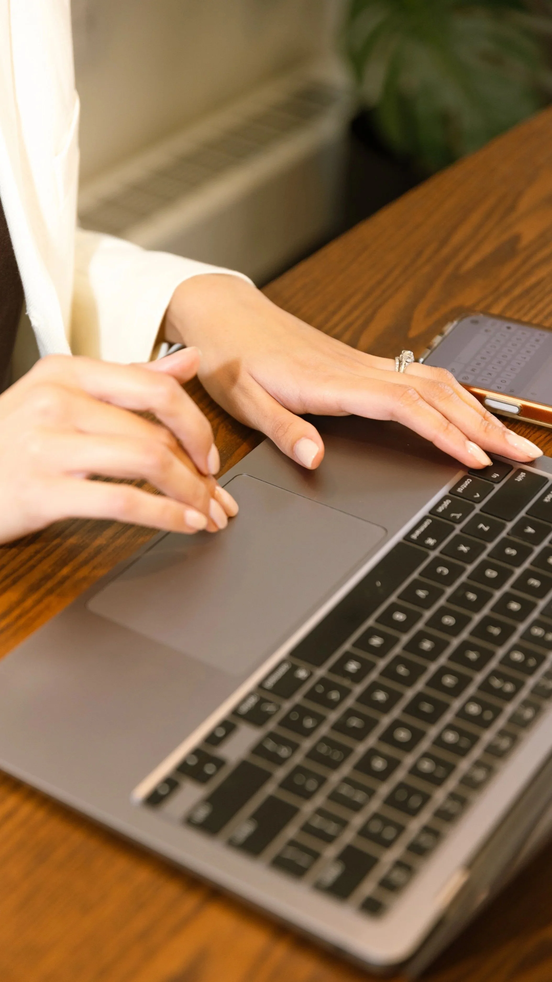 A person typing on a silver laptop with black keyboard, sitting at a wooden desk.