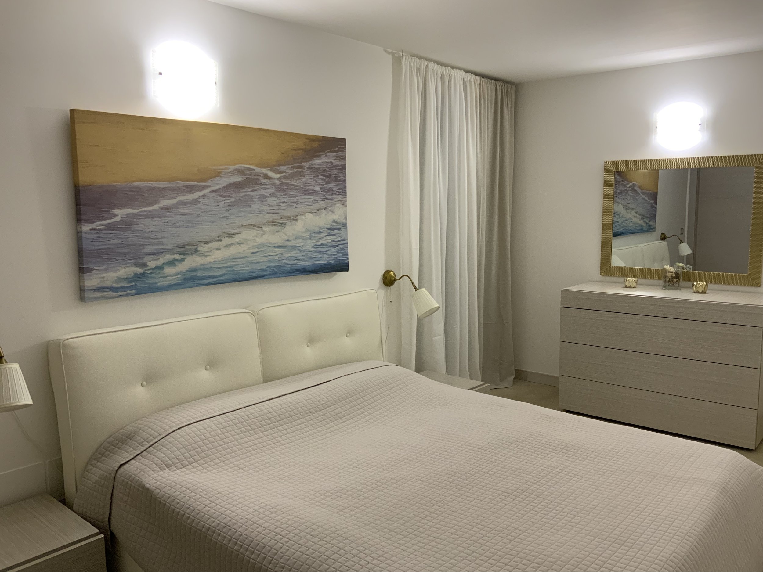 Hotel bedroom with white bed, large ocean wave painting above the headboard, white dresser with mirror, wall-mounted lamps and white curtains.