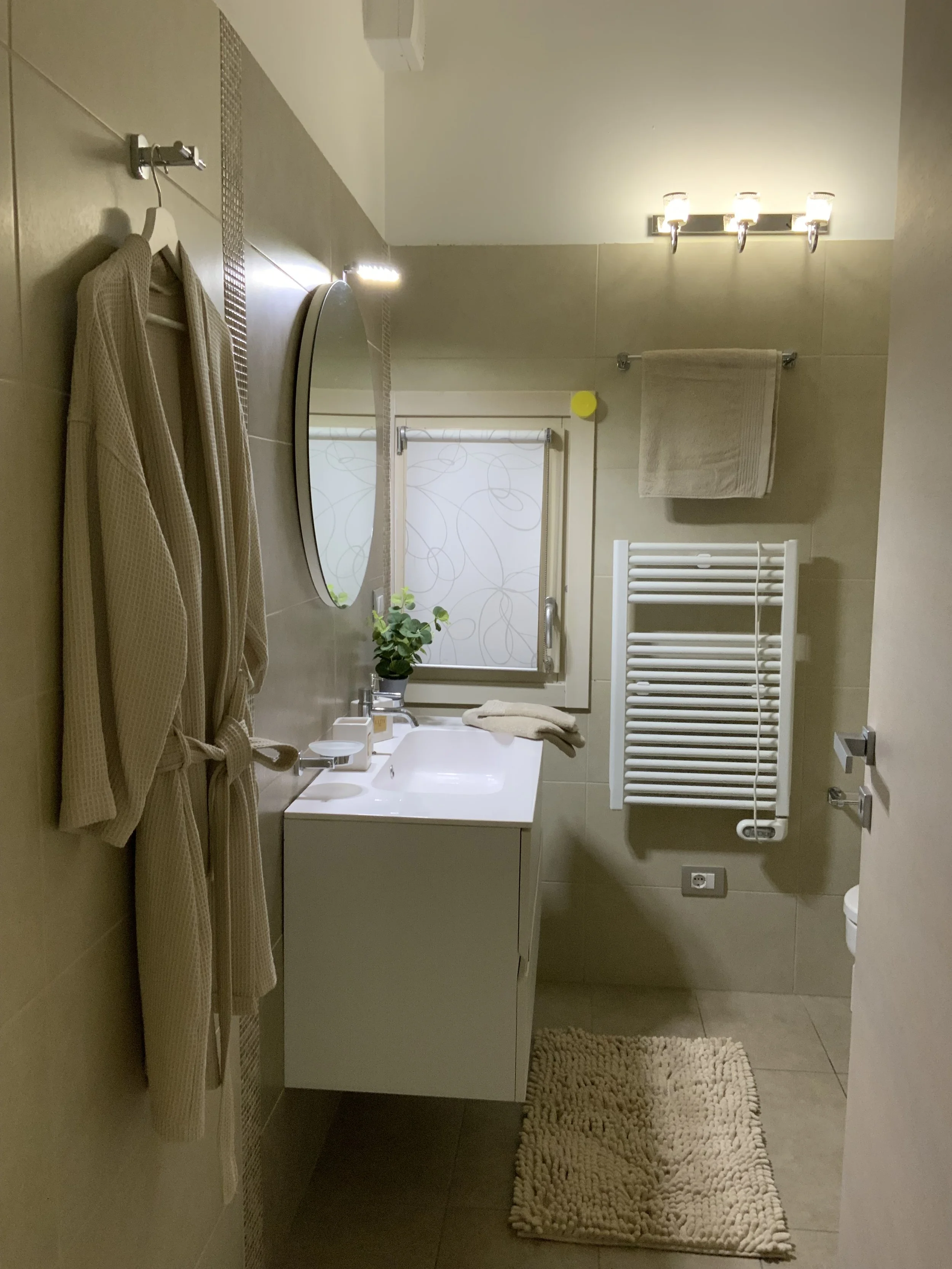 A bathroom with a beige bathrobe hanging on a hook on a tiled wall, a mirror above a white sink, a small potted plant on the window sill, a towel on a rack, a heated towel rail, a rug on the floor, and a window with patterned blinds.