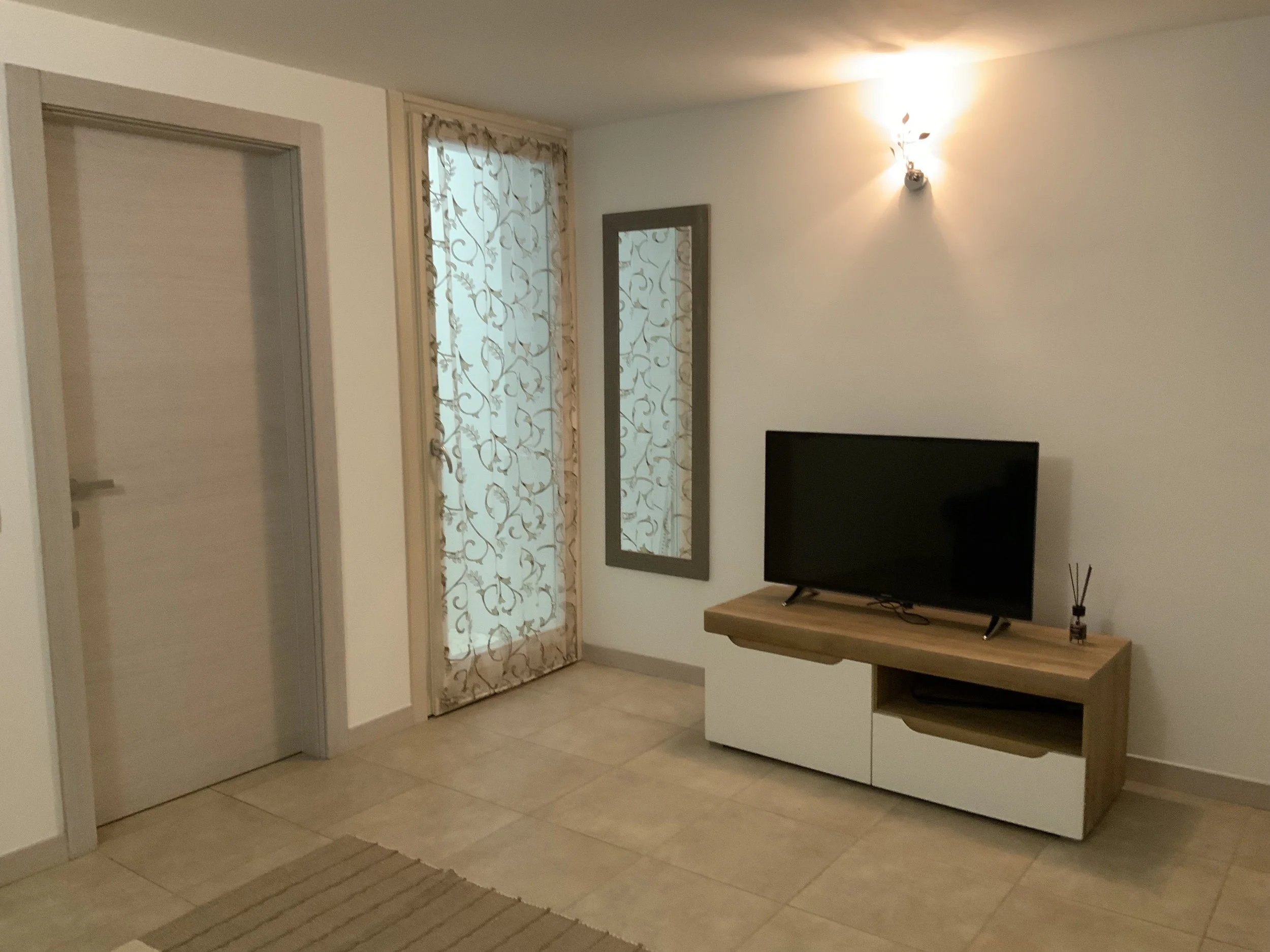 Living room corner with a door, a window with patterned curtains, a mirror, a television on a wooden stand, and a wall light.