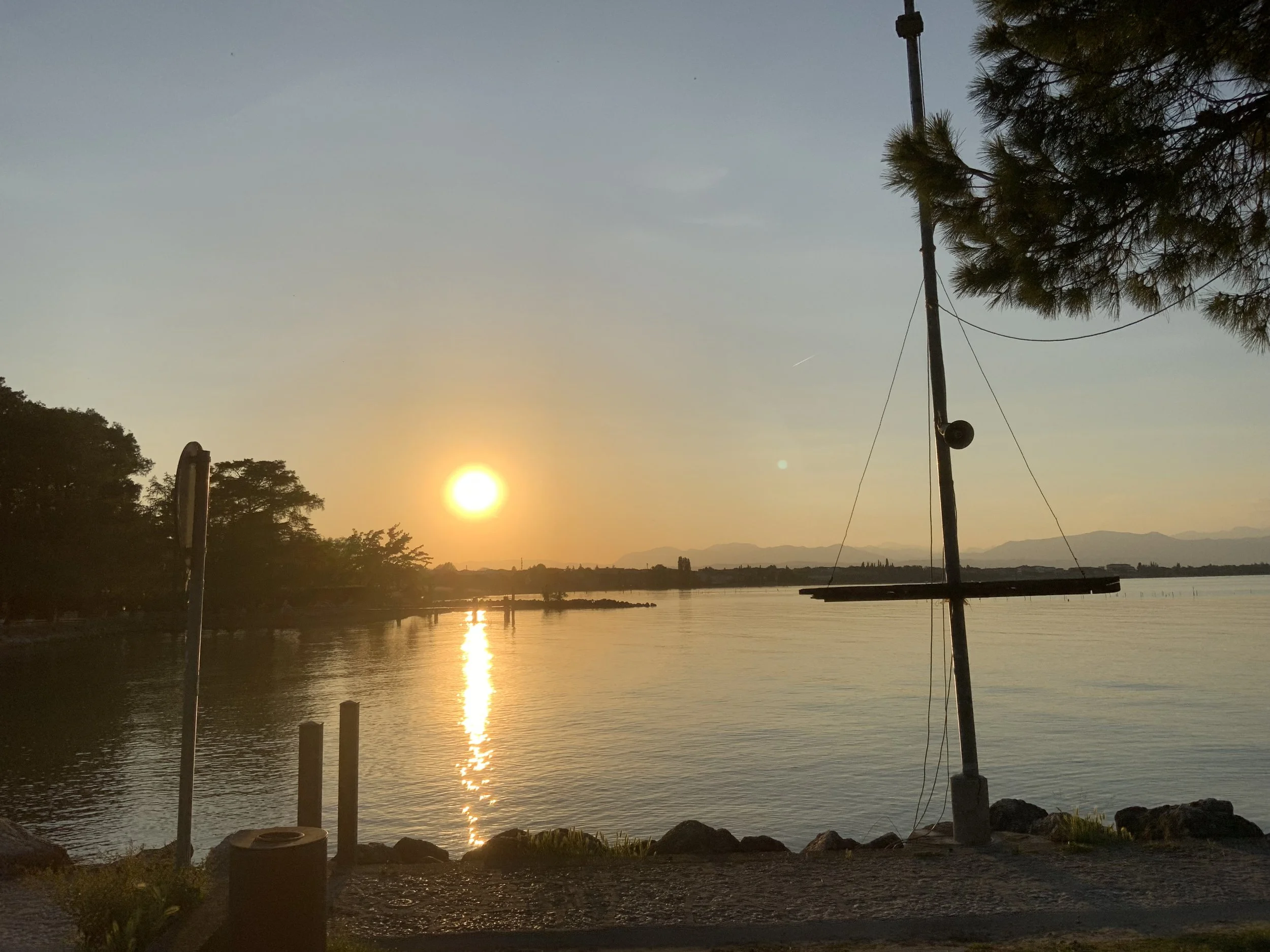 Sunset over a body of water with trees on the left and a sailboat mast on the right, creating a peaceful lakeside scene