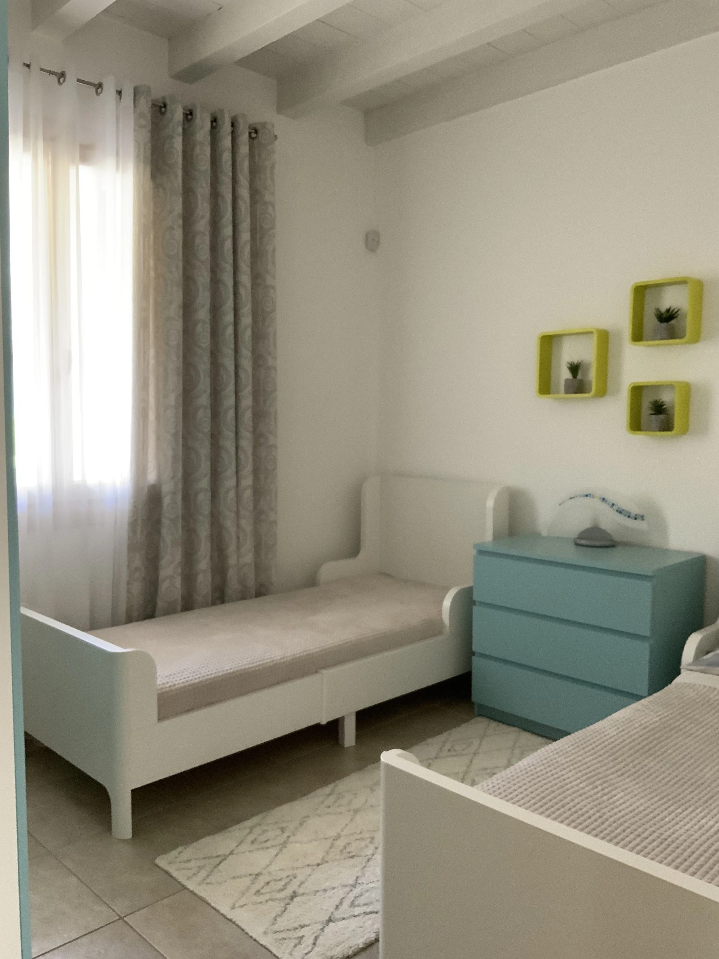 A bedroom with two single beds, white frames, beige mattresses, and striped blankets. A light blue dresser with a gray lamp on top sits between the beds. There are three small green potted plants on yellow wall-mounted shelves to the right of the win