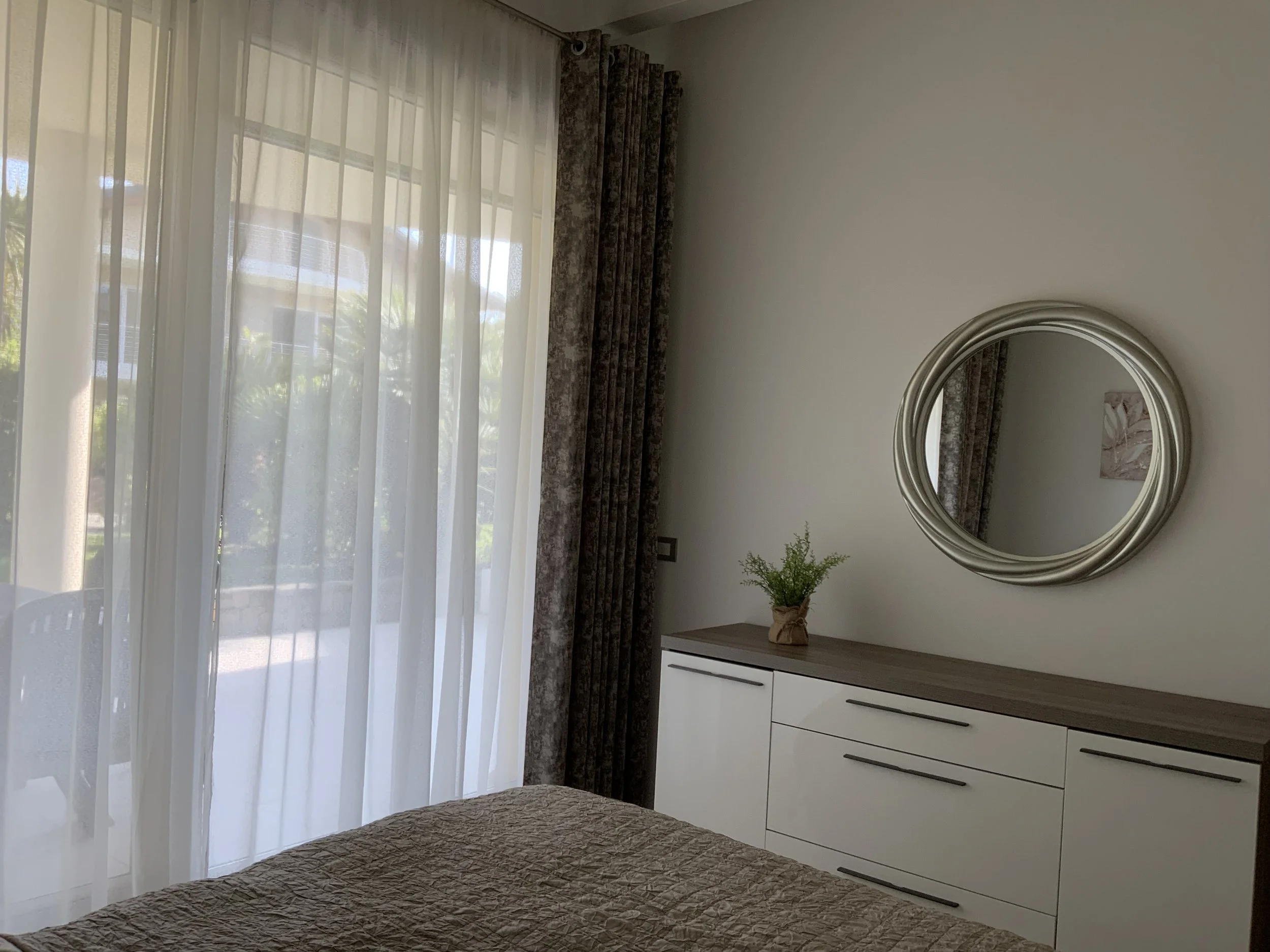 Bedroom with a large window covered by sheer curtains, a white dresser with a wooden top, a round silver mirror on the wall, and a small plant on the dresser.