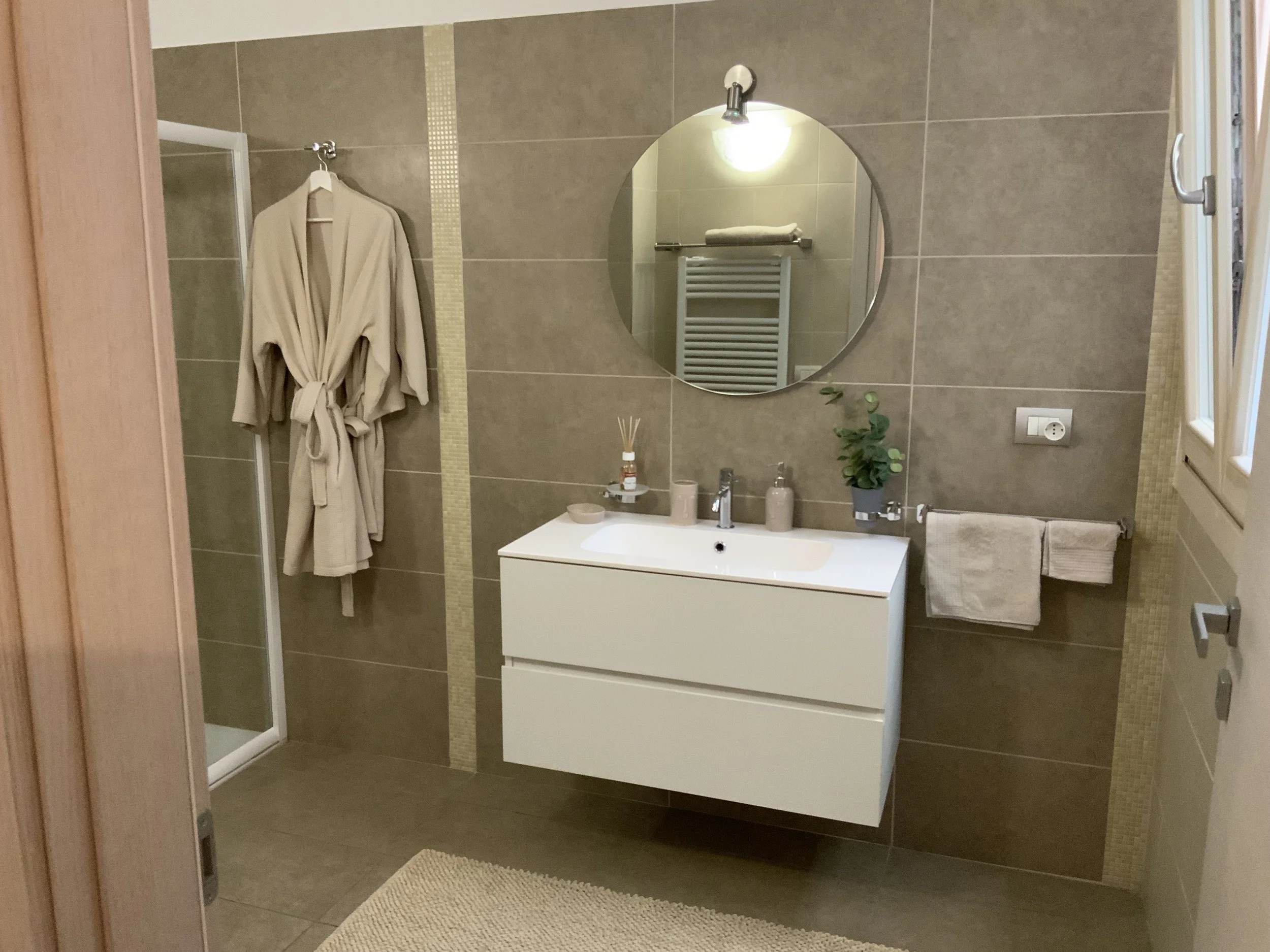 Modern bathroom with beige tiled walls and floor, a white floating vanity with sink, a round mirror, a robe hanging on the wall, and a glass shower enclosure on the left.