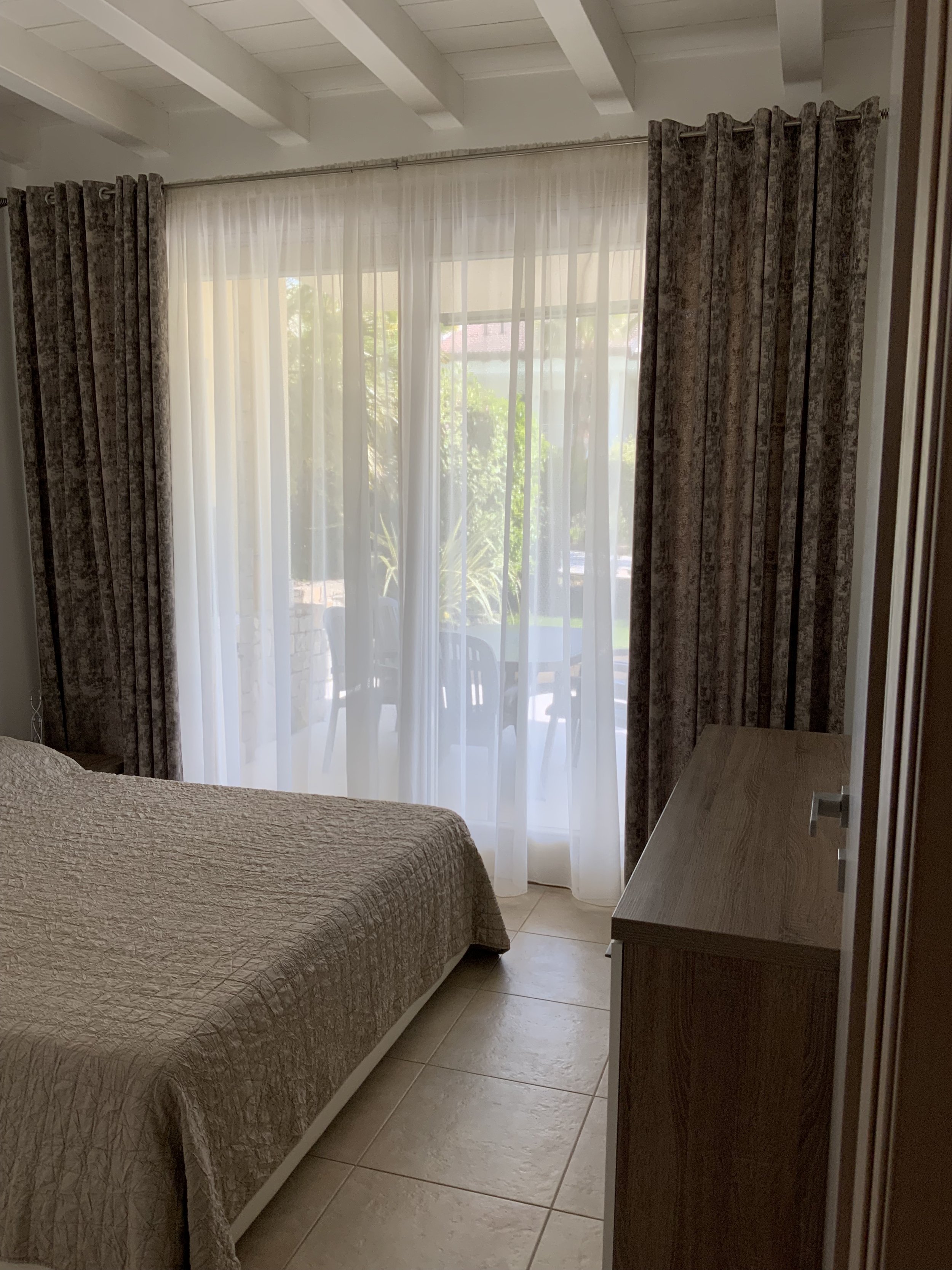 A bedroom with a bed, a wooden dresser, and a sliding glass door covered by sheer white curtains and patterned drapes. There is outdoor patio furniture visible through the glass door and sunlight illuminating the room.