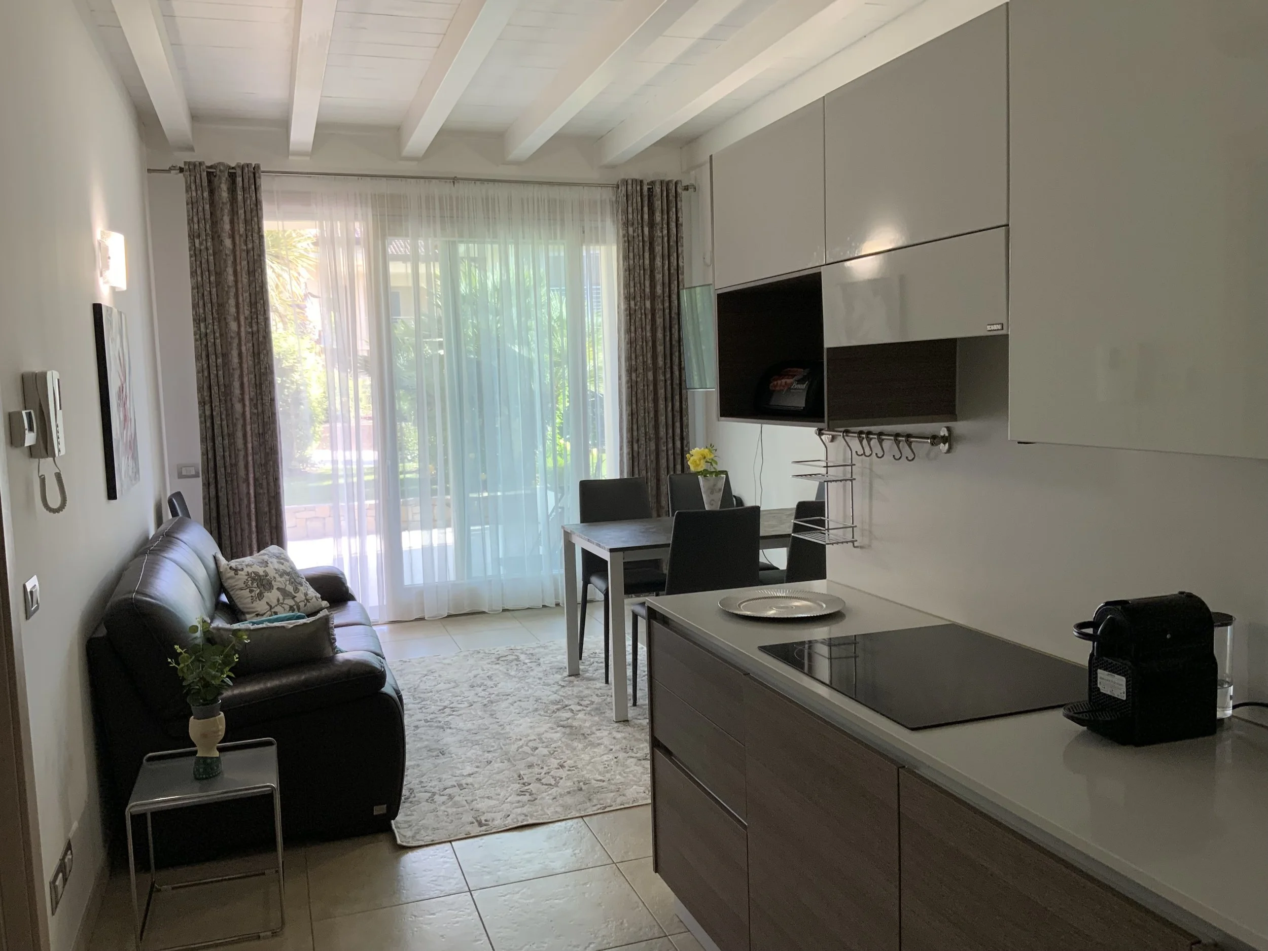 Living room with a sofa, coffee table, dining table with chairs, and kitchen area with stove and coffee machine, large glass doors with sheer curtains revealing outdoor greenery.
