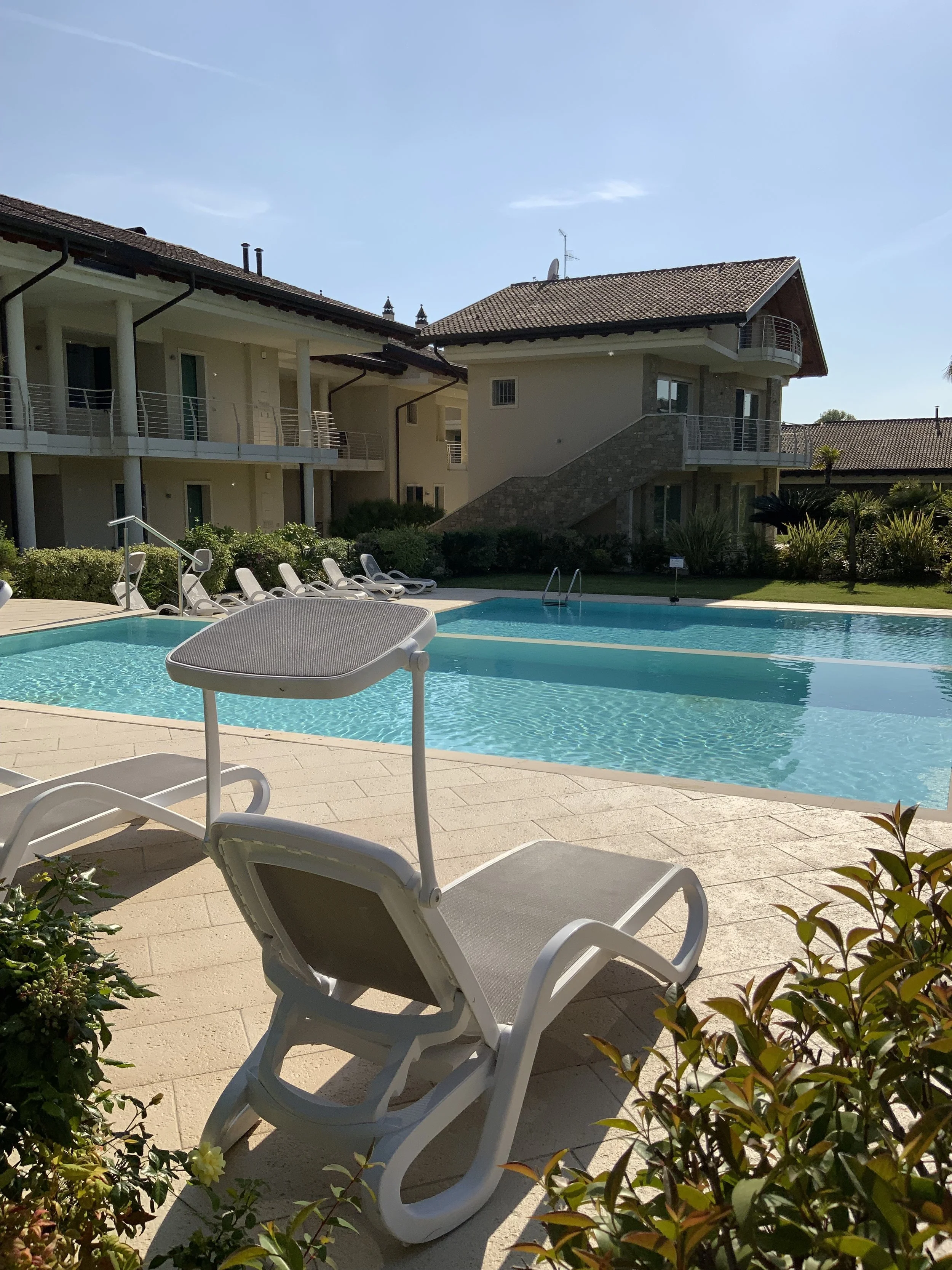 Swimming pool with lounge chairs and a shaded seat, surrounded by low bushes, in a residential area with apartment buildings and houses, under a clear blue sky.