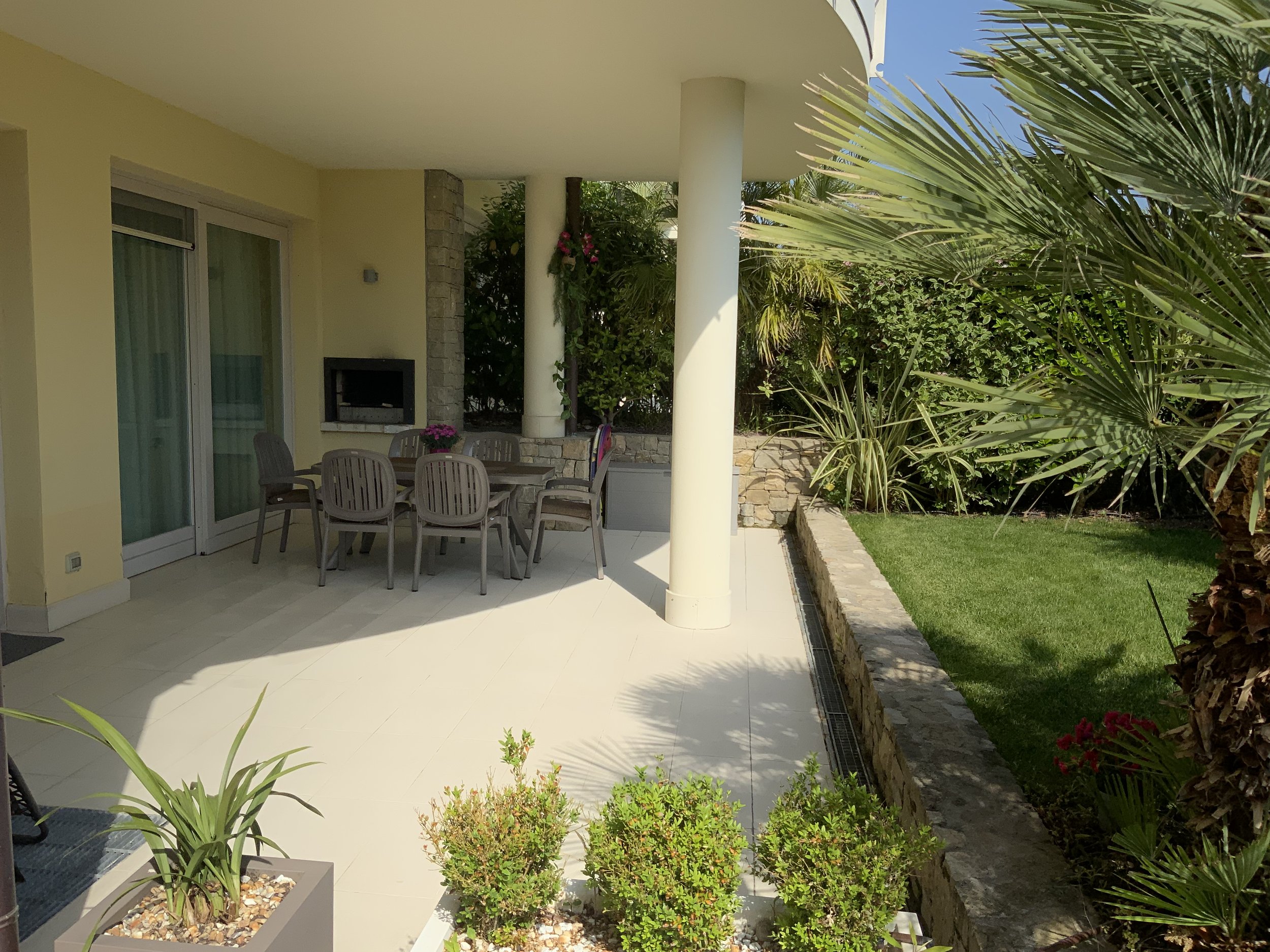 Outdoor patio area with a round dining table and six chairs, a built-in grill, a sliding glass door, and a lawn with lush green plants and trees.